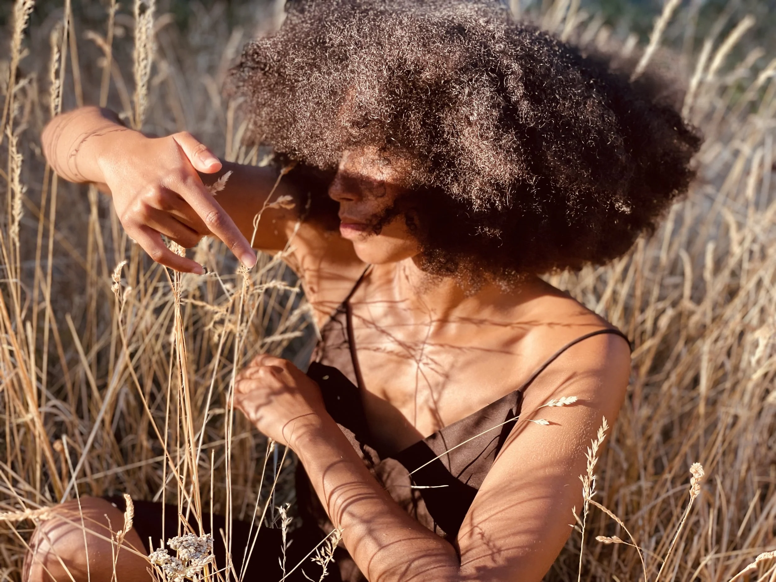 A woman with curly hair sitting in a field of dry, tall grass, wearing a black top, and making a peace sign with her hand.