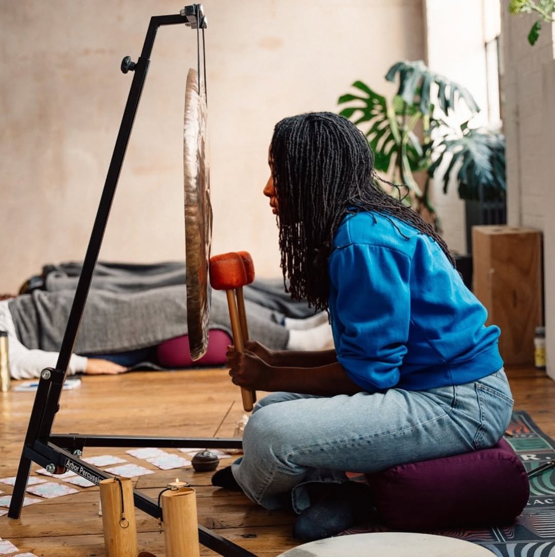 A person with long braided hair sits on a cushion and looks at a large gong hanging from a black stand, holding a mallet. The setting is a cozy indoor space with a wooden floor, some houseplants, and a person lying on a couch in the background.