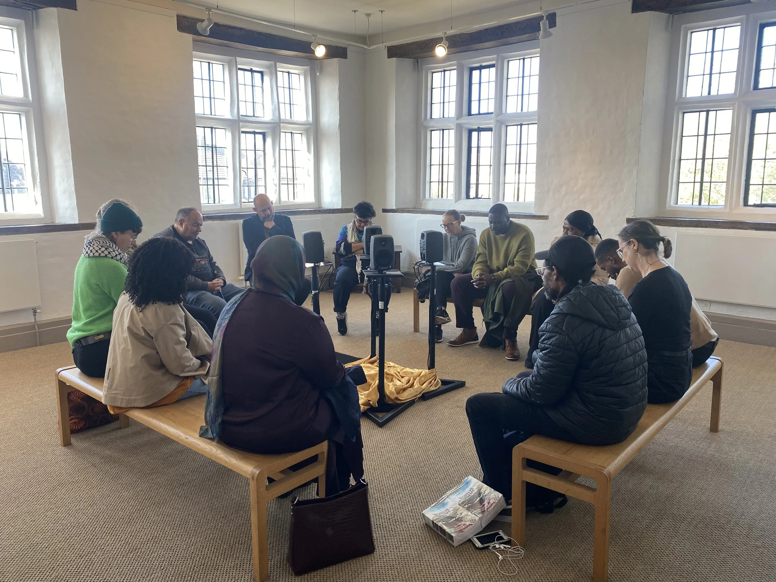 People sitting in a circle in a bright room with large windows, participating in a group discussion or meeting, with audio equipment and personal belongings on benches.