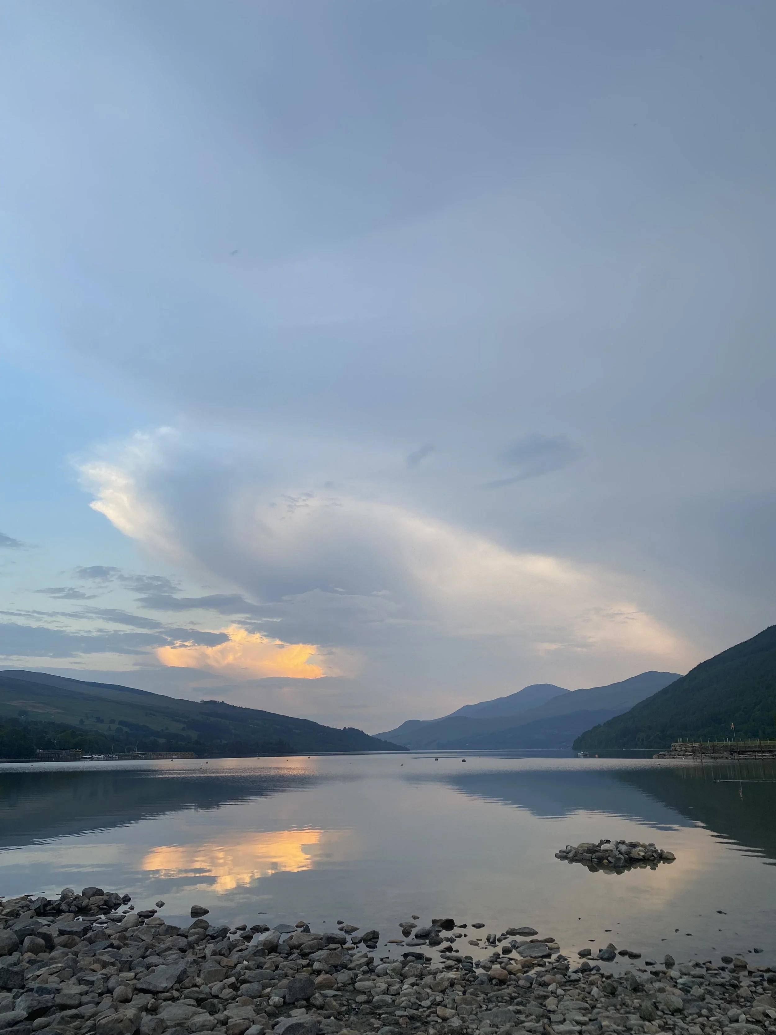 A scenic view of a lake surrounded by mountains with a cloudy sky at sunset, with the water reflecting the sky and landscape.