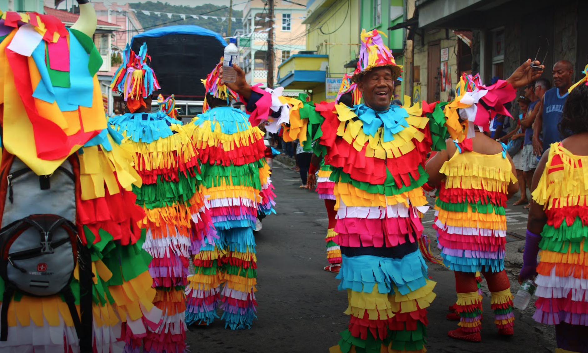People dressed in colorful, fringed costumes participating in a street carnival or festival parade, with street buildings and onlookers in the background.