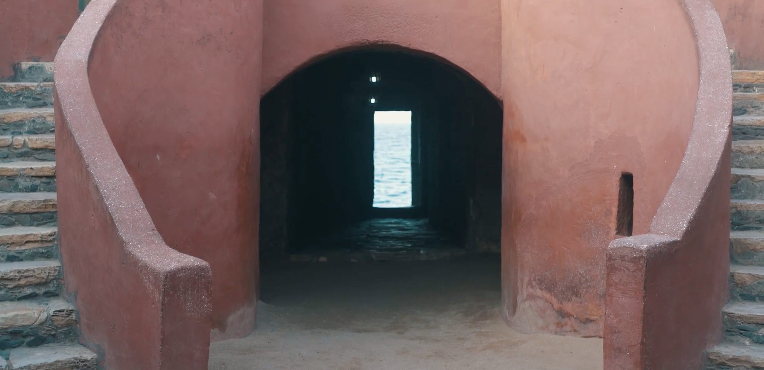 A tunnel built into a reddish structure with steps on either side, leading to a view of water through the tunnel opening.