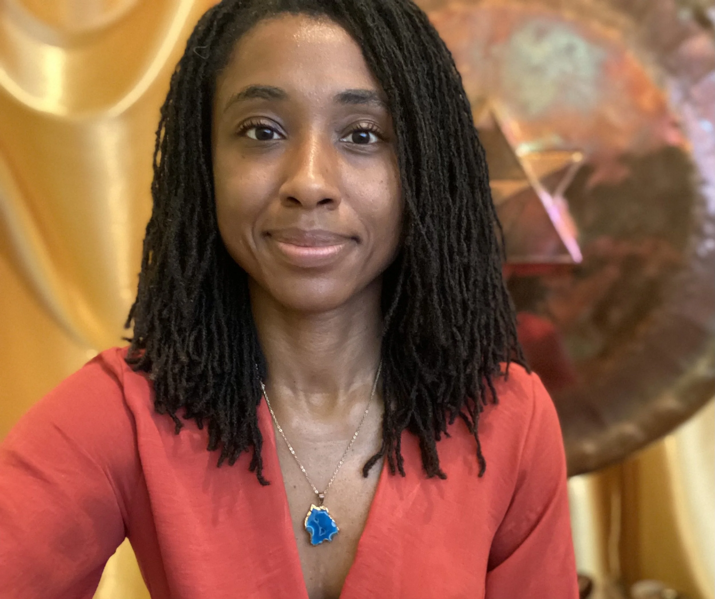 Close-up of a woman with long dreadlocks and wearing a red top, smiling softly. She is wearing a blue pendant necklace and a silver chain. The background features warm tones with yellow and reddish-orange abstract patterns.
