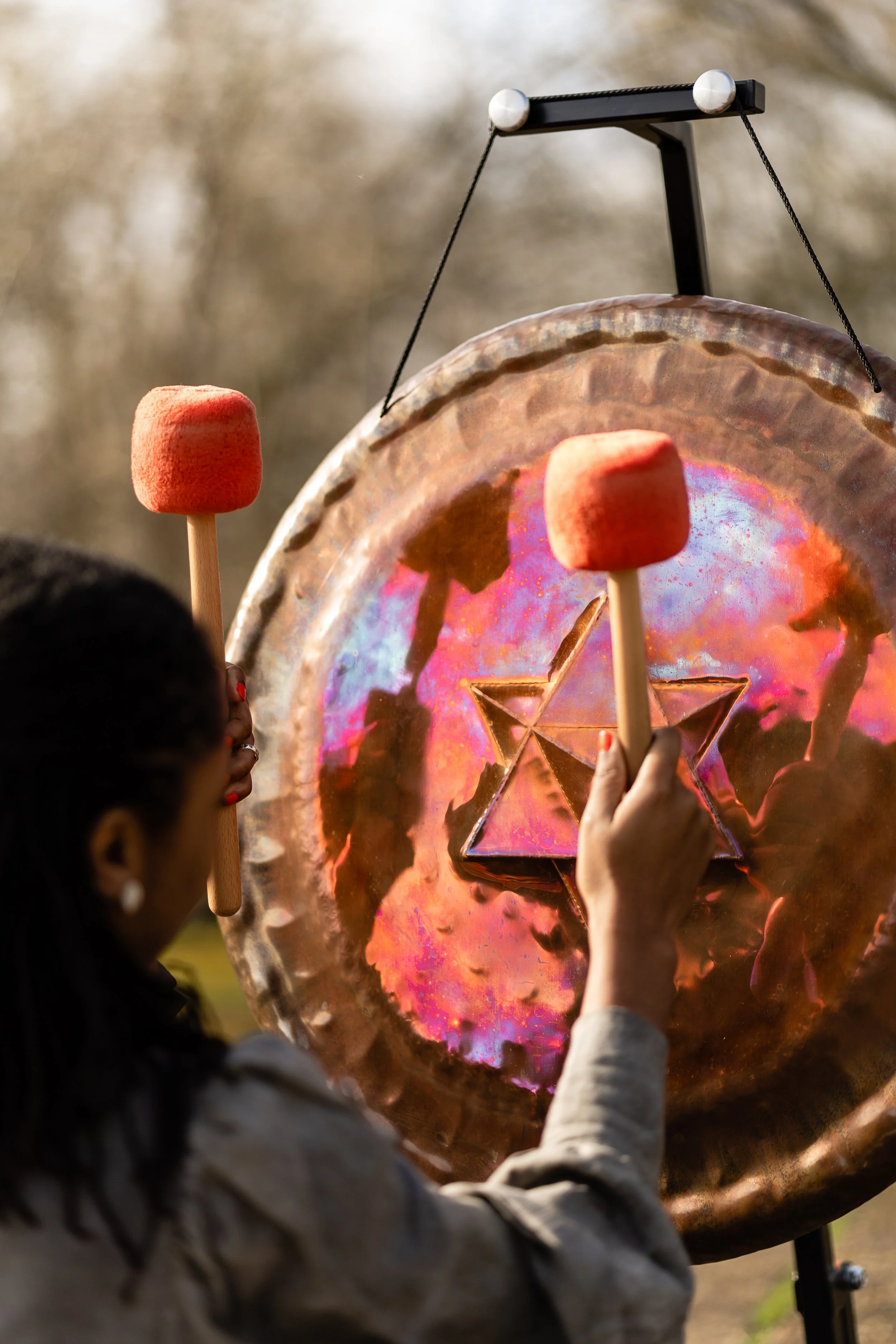 A person is striking a large gong with a drumstick topped with a red felt pad. The gong has a metallic, iridescent surface with a star-shaped design engraved in the center.