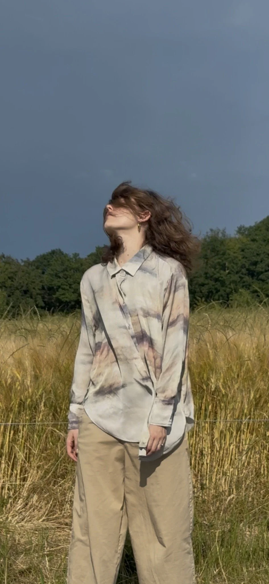 A woman standing in a field of tall grass with a dark stormy sky in the background, her hair blowing in the wind.