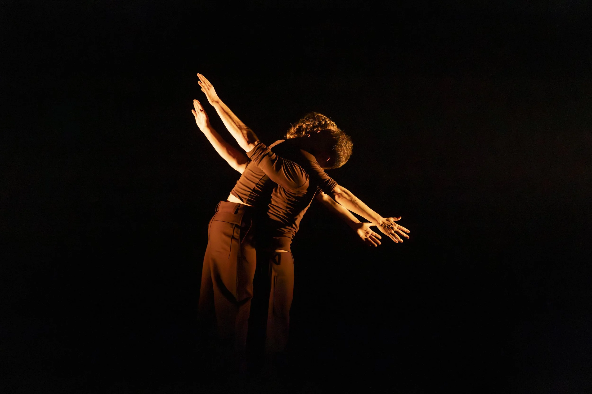 Three dancers performing on stage in a dark environment, with their arms extended outward and downward, illuminated by warm lighting.