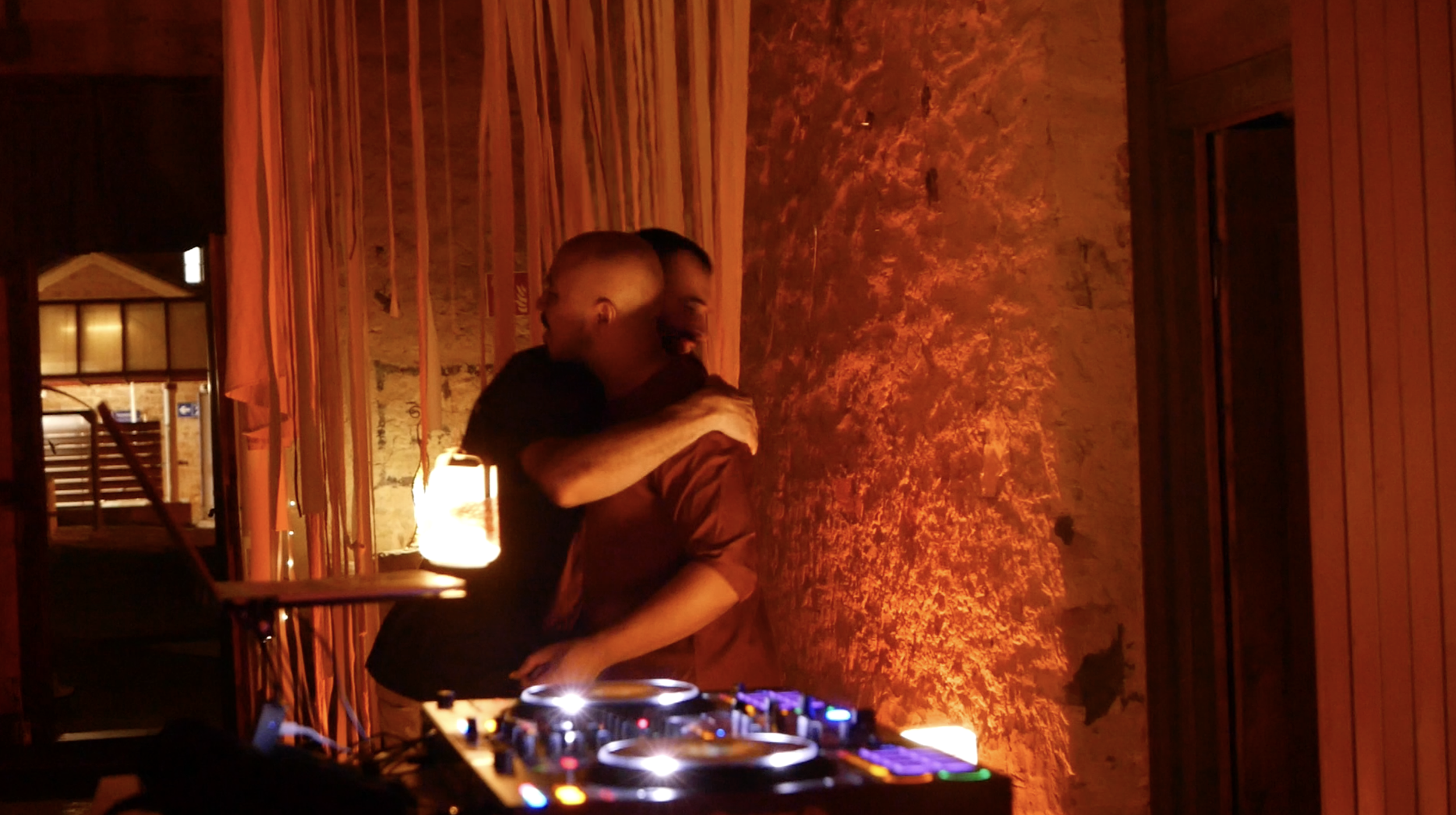 Two people embracing in a warmly lit room with wooden walls and decor, standing near a DJ setup with colorful lights.