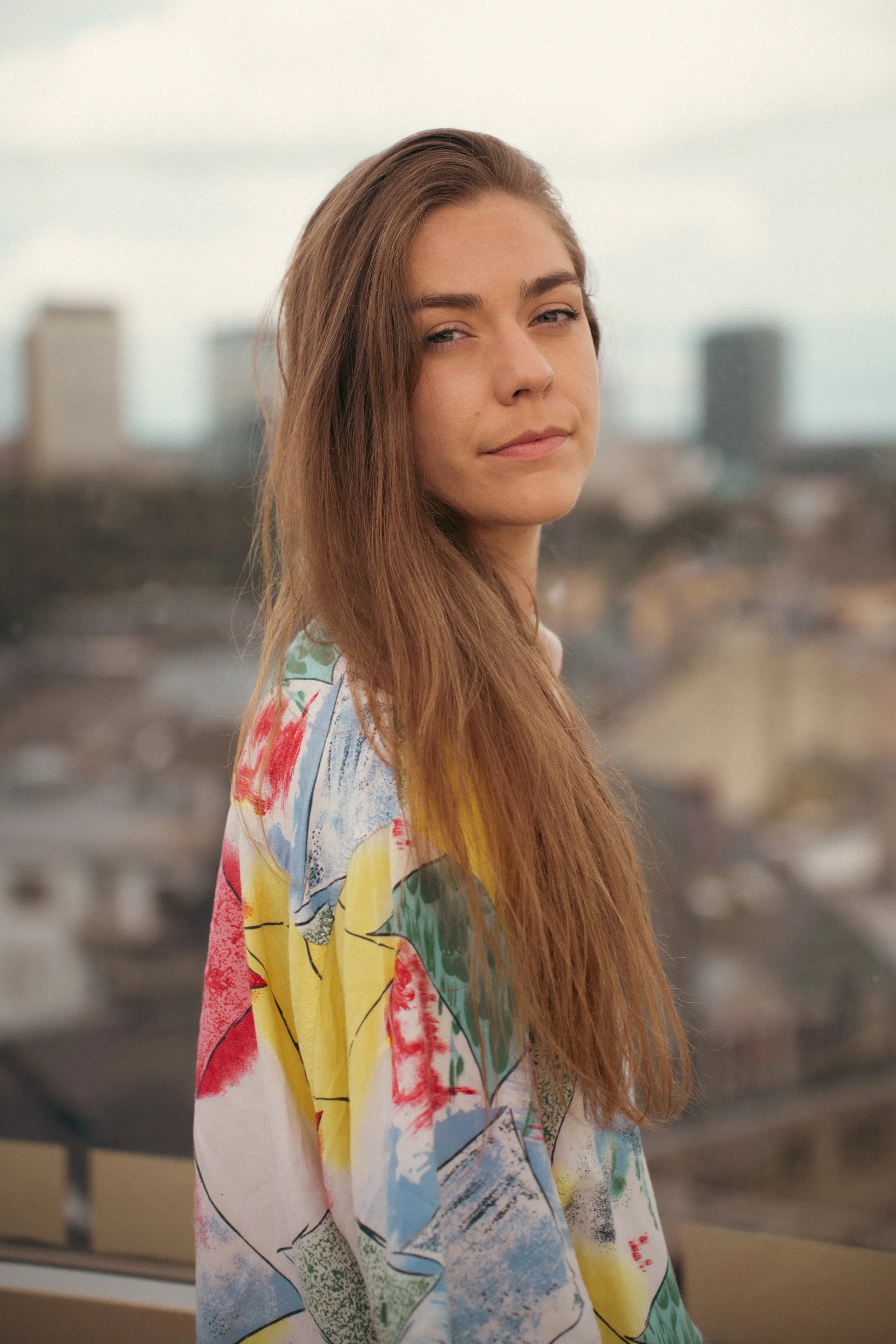 A young woman with long, light brown hair and fair skin standing outdoors on a cloudy day with a cityscape in the background. She is wearing a colorful, abstract-patterned blouse.