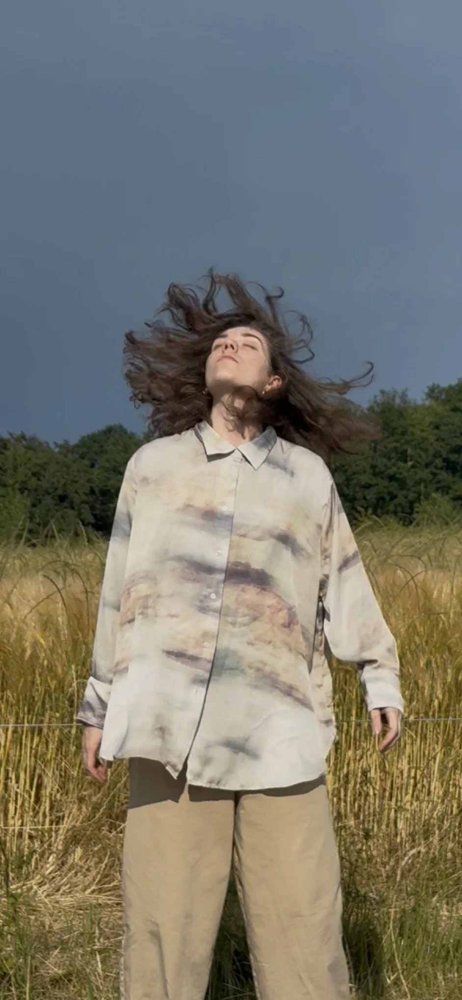 A woman standing in a field of tall grass with her hair blowing in the wind, wearing a loose, patterned shirt and beige pants, with a dark stormy sky in the background.