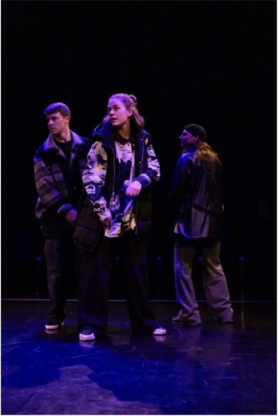 Three young adults standing on stage in dim lighting, with dark background, appearing to perform or rehearse.