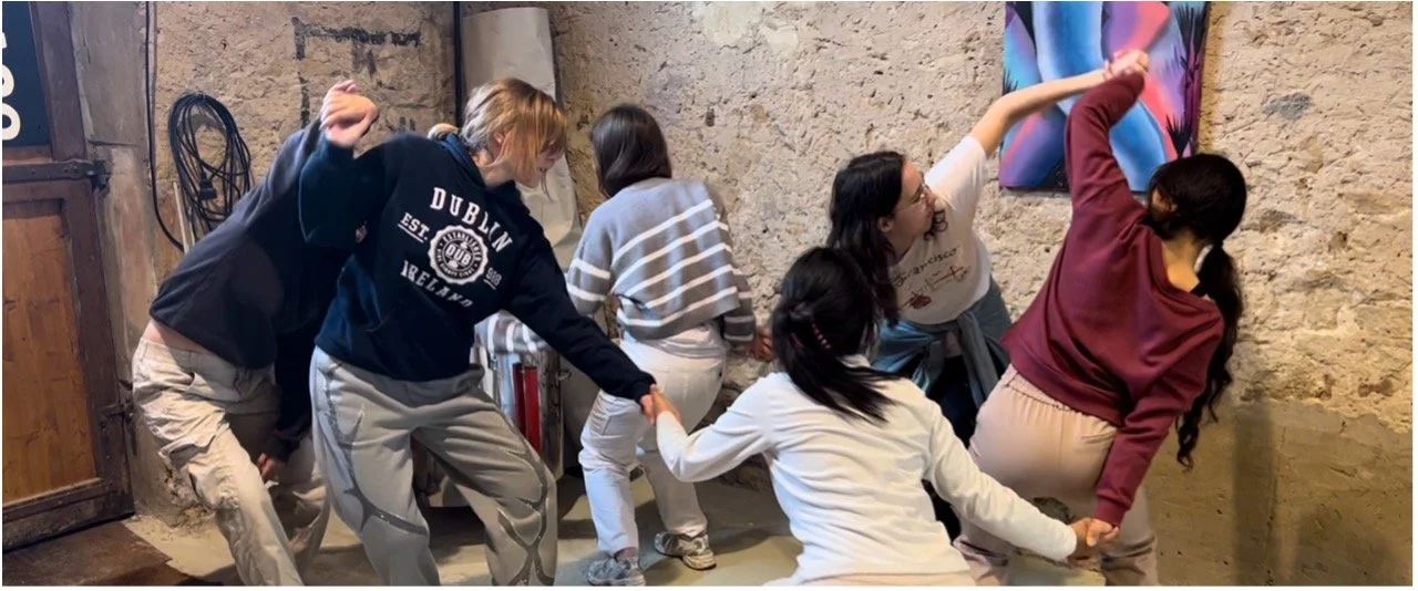 Five women playing a game involving holding hands and leaning back in a room with stone wall. One woman is wearing a gray striped sweater, another in a white shirt, another in a maroon hoodie, and the others in casual clothing. They are smiling and having fun.