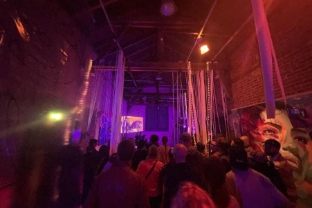 Audience at an indoor concert or event with colorful lighting, hanging decorations, and a stage with visuals in the background.