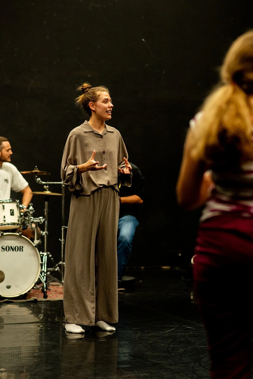 Woman speaking or singing on stage with a band, including a drummer in the background, in a black rehearsal space.