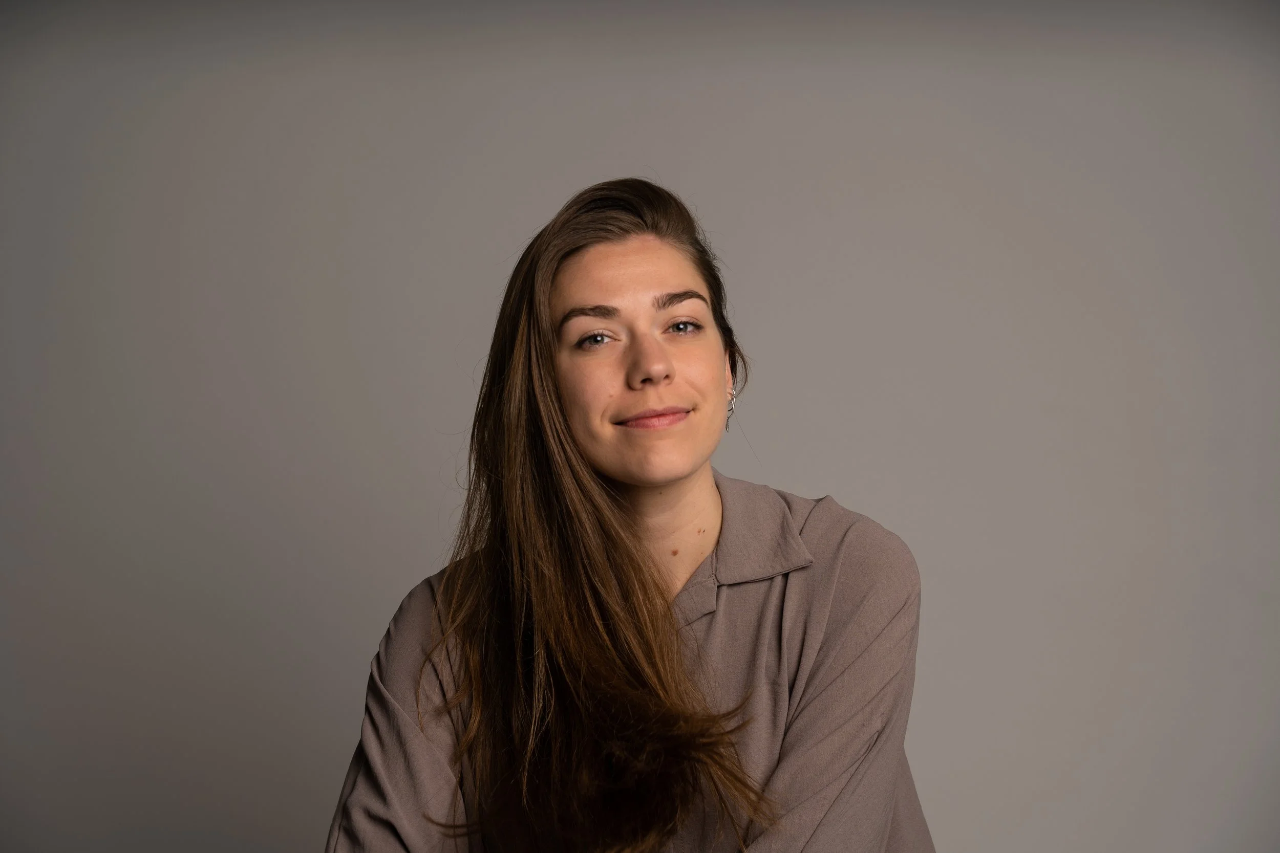 A woman with long brown hair wearing a taupe shirt, looking at the camera with a slight smile, against a plain gray background.