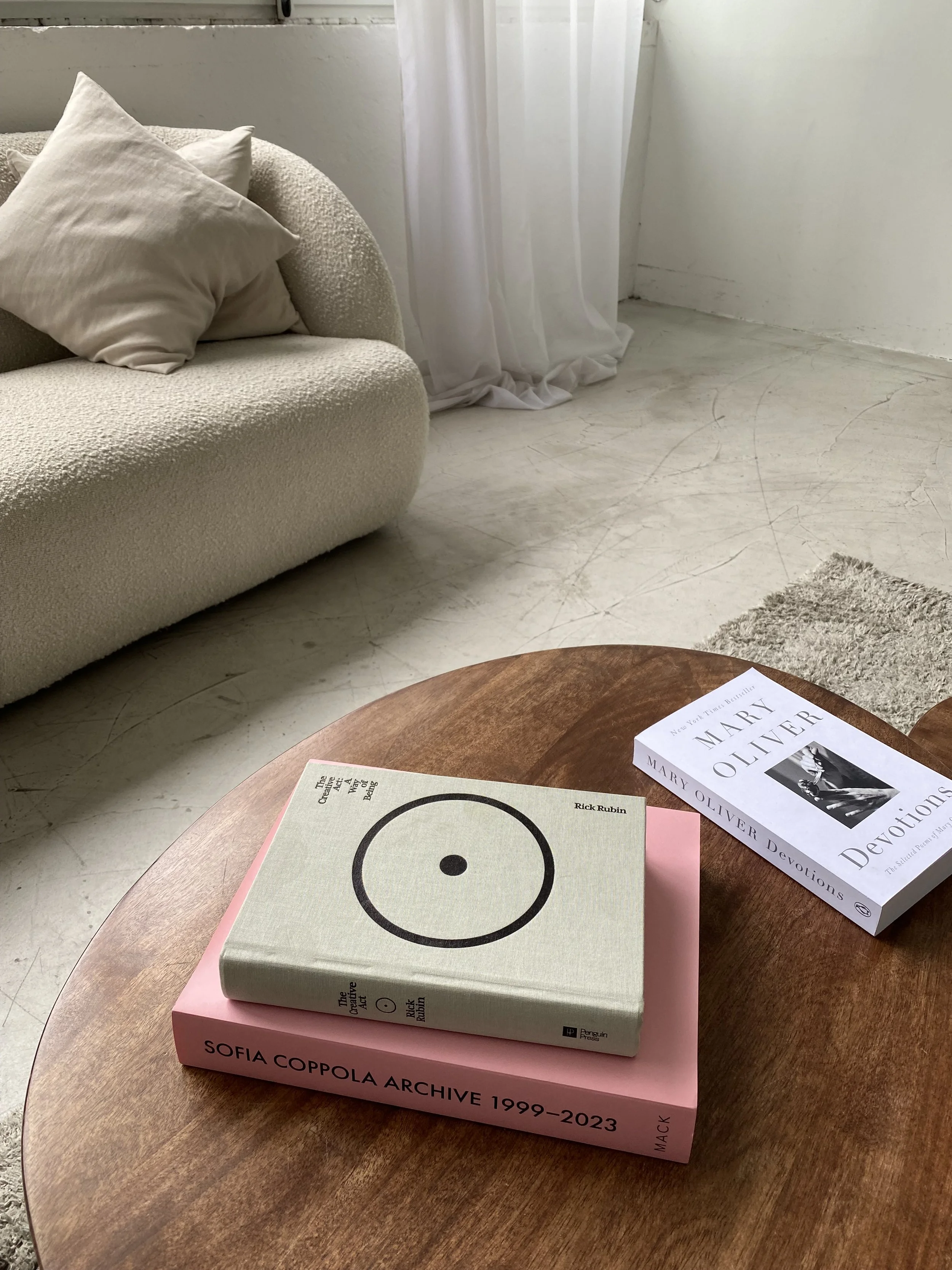 Ein runder Holztisch mit drei Büchern darauf. Das oberste Buch ist grau mit einem schwarzen Yin-Yang-Symbol. Darunter liegt ein pinkfarbenes Buch mit dem Titel "Sofia Coppola Archive 1999-2023". Im Hintergrund ein Wohnzimmer mit weißen Wänden, einem cremefarbenen Sofa mit Kissen, weißen Vorhängen und einem hellen Boden.