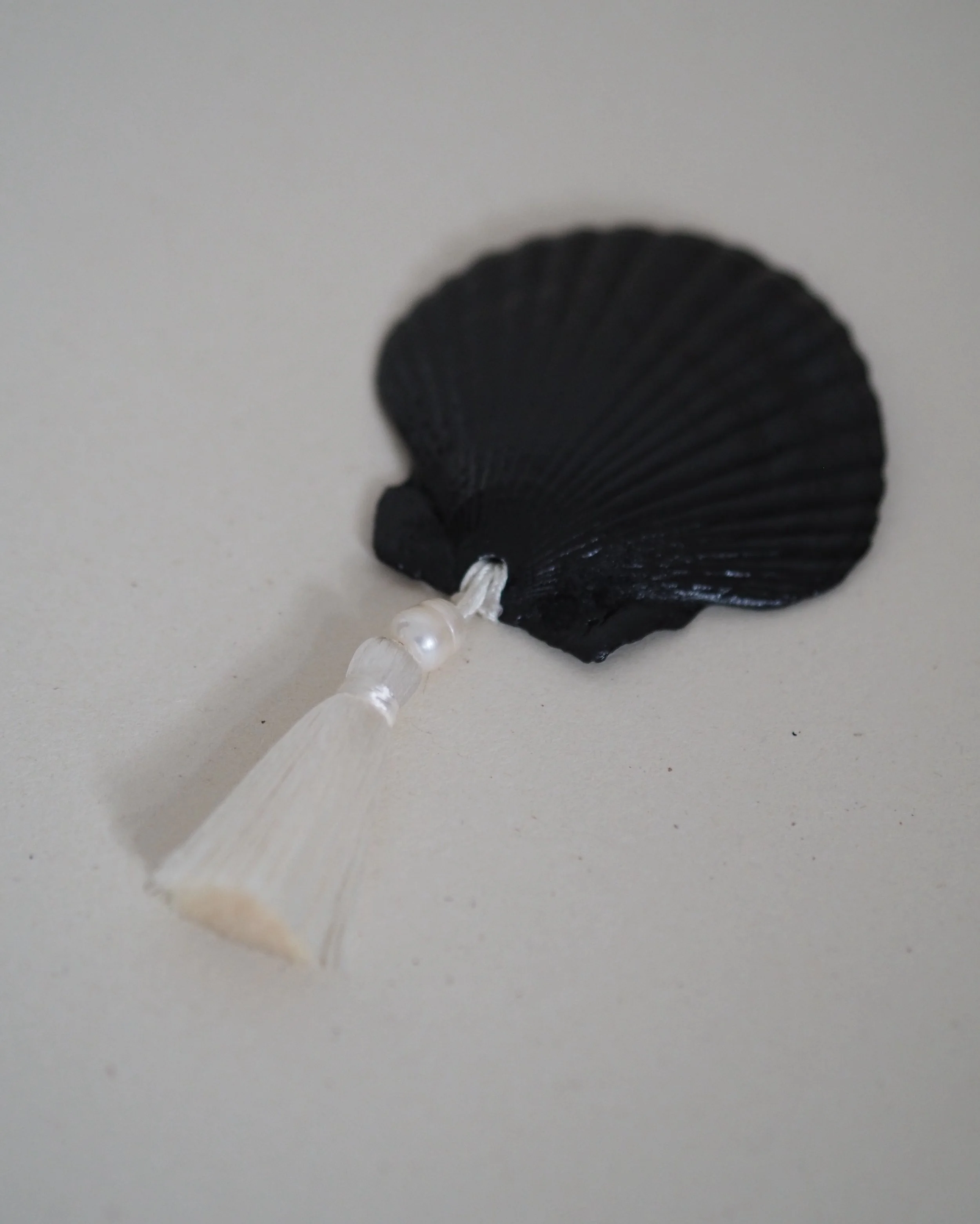 Eine schwarze Muschelschale mit weißen Perlchen, befestigt an einem weißen Band, liegt auf einer flachen, hellgrauen Oberfläche.