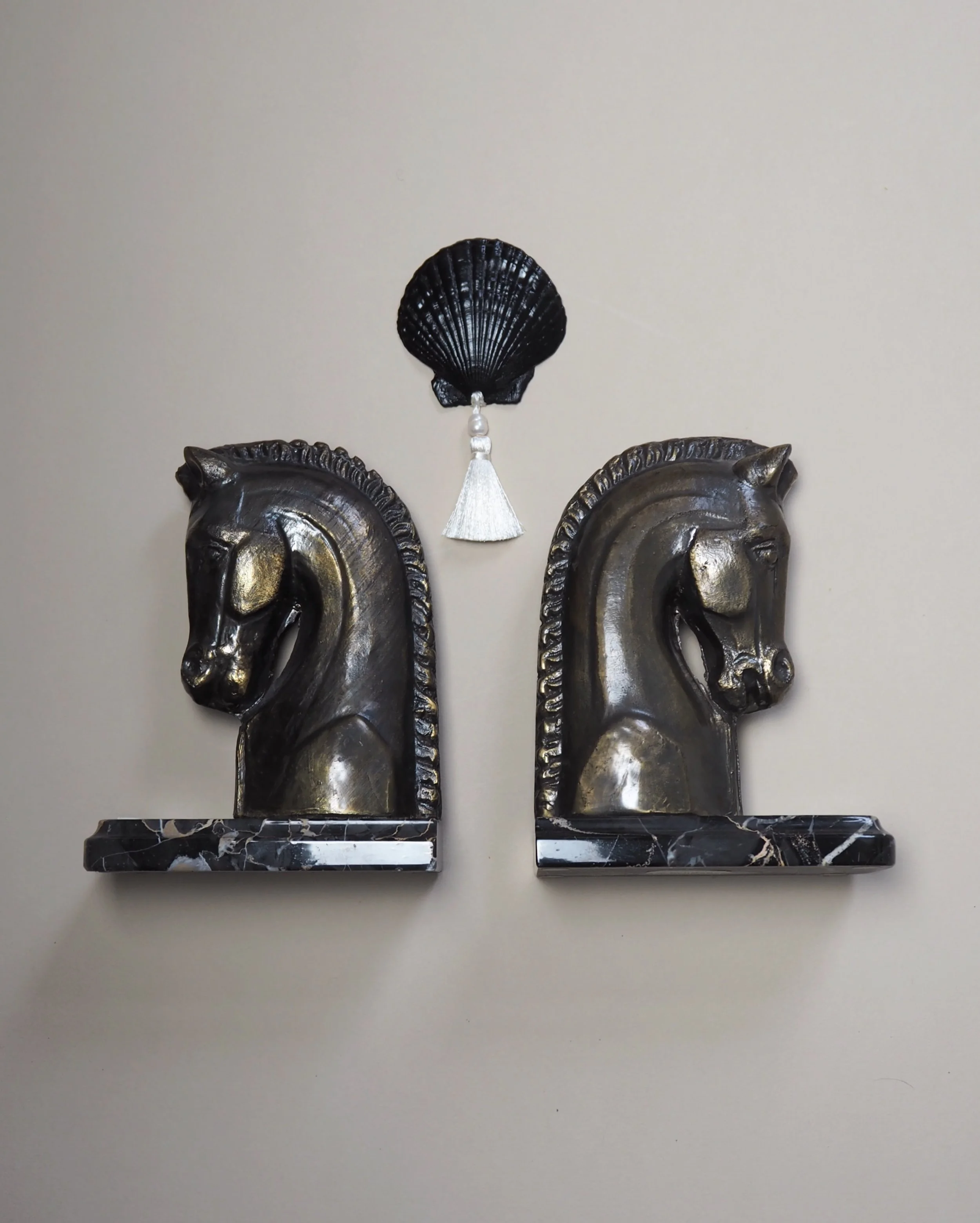 Zwei silberne Wanduhren mit dunklem Rahmen stehen auf einem schwarzen Marmorsockel, darüber hängt eine schwarze Muschelschale mit einer weißen Quaste. Die Wand im Hintergrund ist hell glatt.