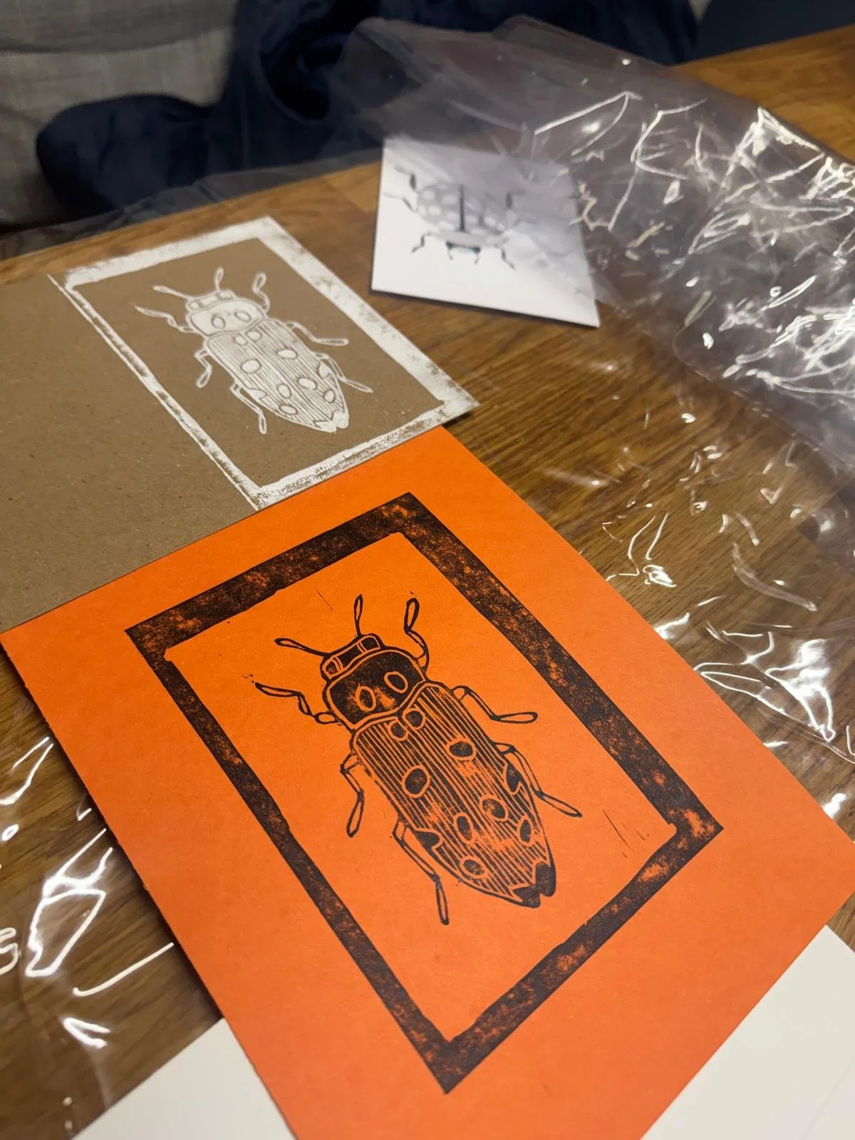 Printed illustration of a beetle on various sheets of paper, some with a distressed border or background, placed on a wooden surface with clear plastic wrap nearby.
