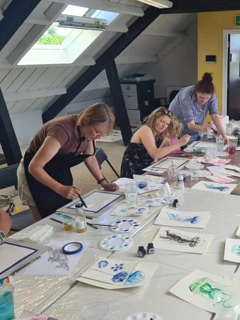 People participating in a watercolor painting workshop in a bright room with sloped windows, multiple paint palettes, brushes, and artwork on the table.
