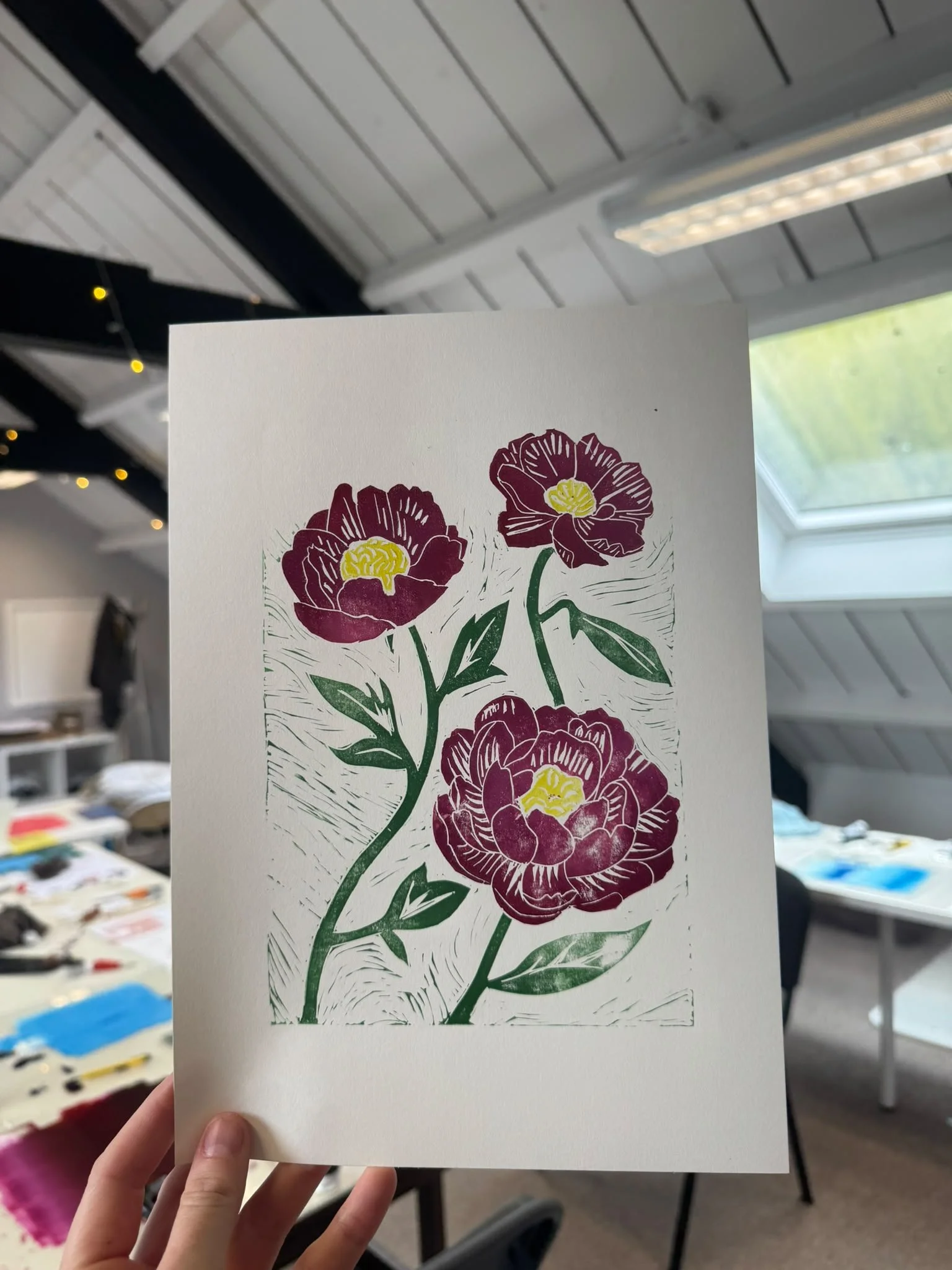 A hand holding a sheet with a colorful floral illustration of maroon, yellow, and green flowers and leaves, against a background of a room with a ceiling, window, and craft supplies.
