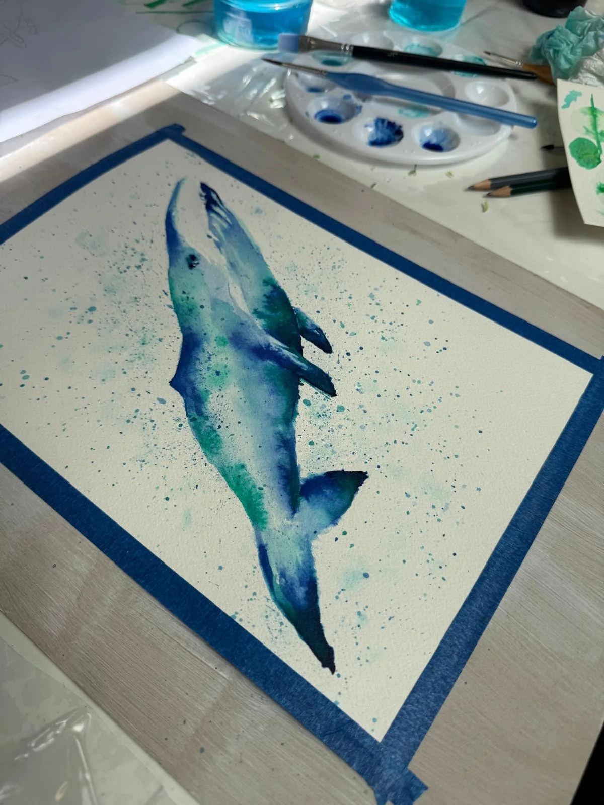 Watercolor painting of a fish on paper with blue and green hues, surrounded by paint splatters. Art supplies, including brushes, a paint palette, and tissue paper, are visible on the table.