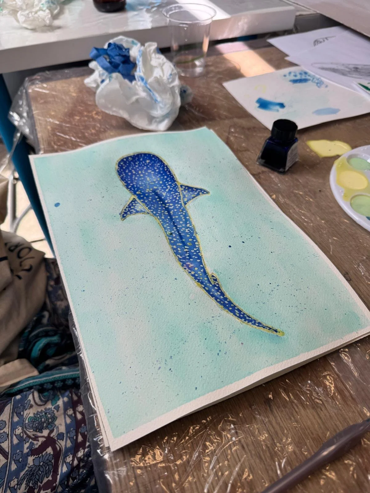 A watercolor painting of a blue whale with yellow dots on a light background, sitting on a table alongside art supplies including watercolor paints, paper, and a water cup in an indoor art space.