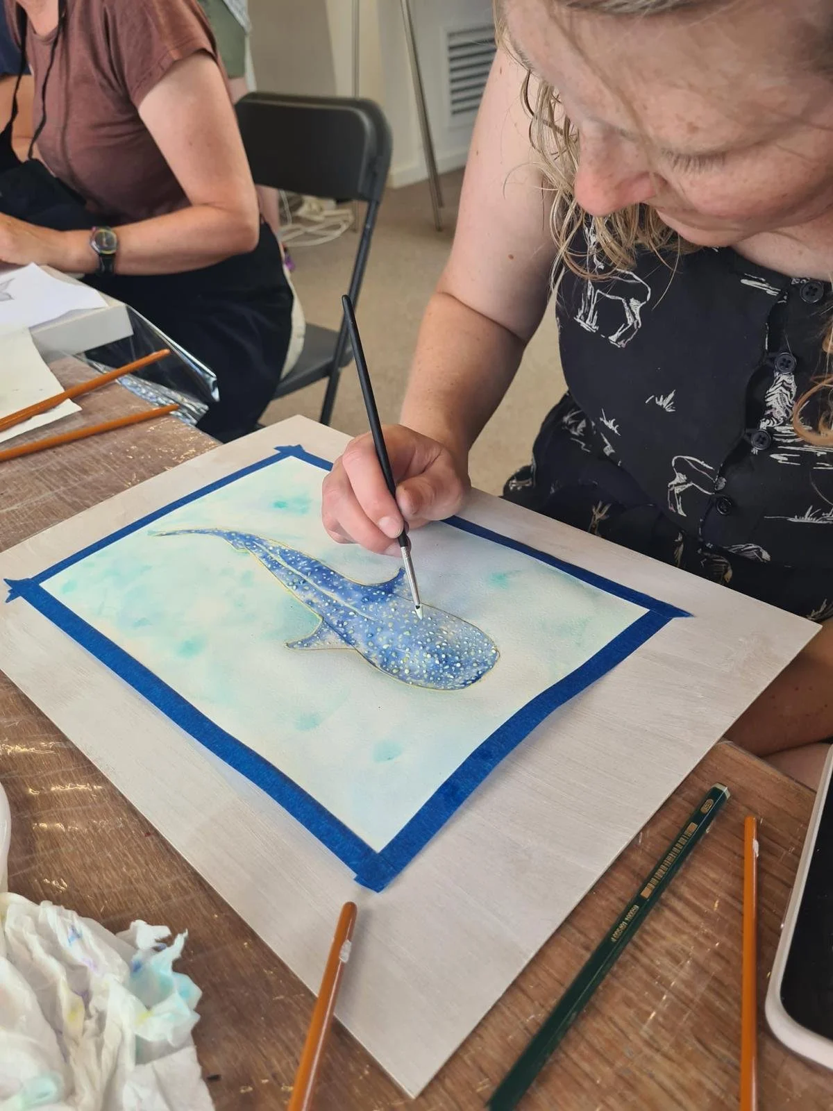 Woman painting a watercolor picture of a whale on a piece of paper, with various art supplies on a wooden table.