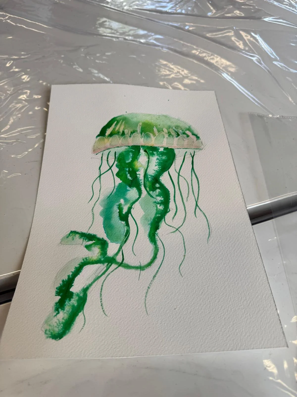 A watercolor painting of a jellyfish with green, pink, and white hues on textured watercolor paper.