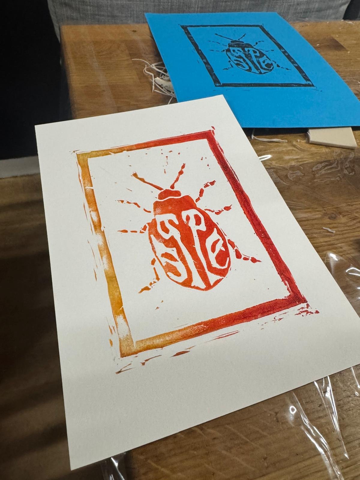Wooden table with two pieces of paper featuring a stencil-style bug design, one in orange and one in blue.