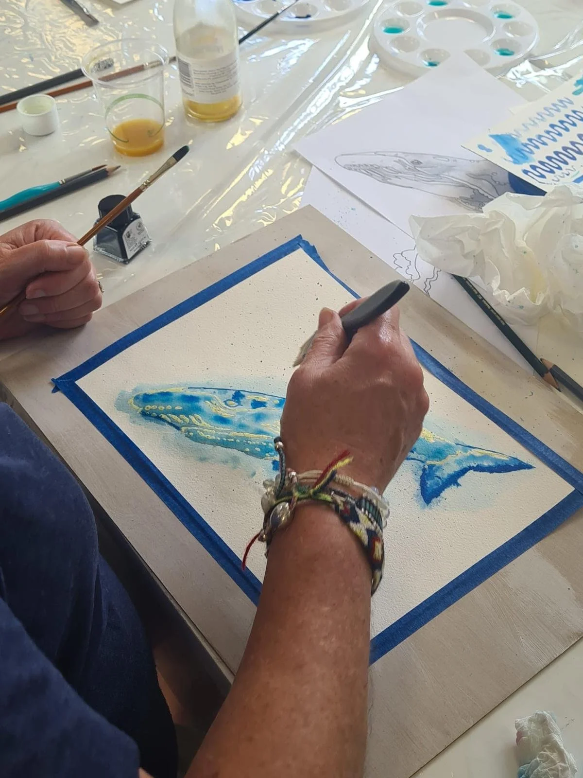 Person creating a watercolor painting of a whale on a piece of paper, surrounded by painting supplies.