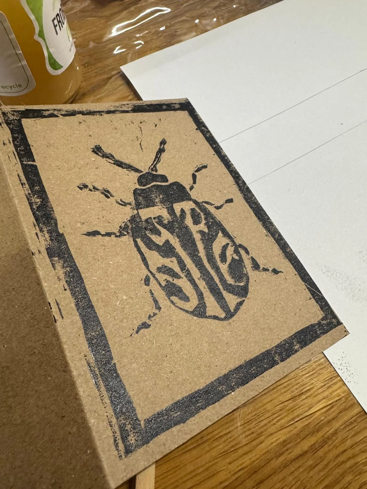 A cardboard box with a stamped image of a beetle on its lid, placed on a wooden table with a piece of white paper and a beverage bottle nearby.