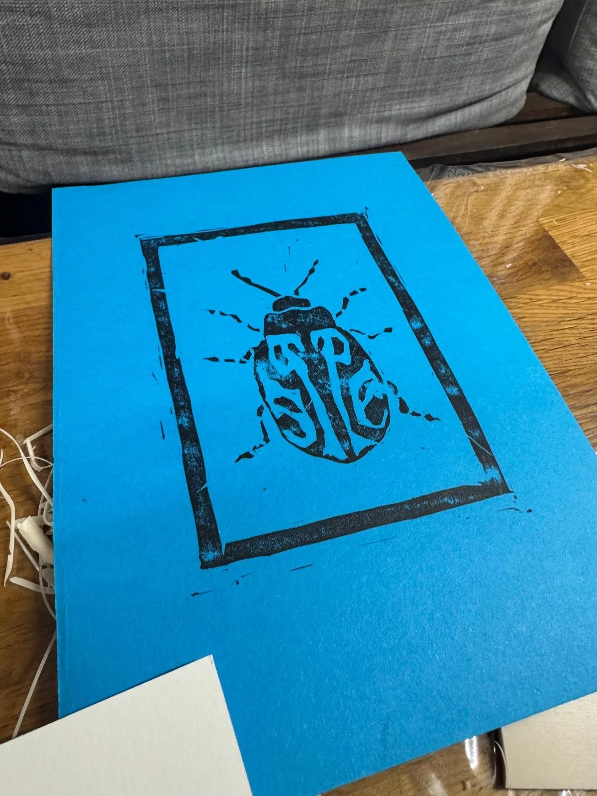 Blue paper with a black stencil of a bug with the letters S, P, A, C inside it, framed by a rectangular border.
