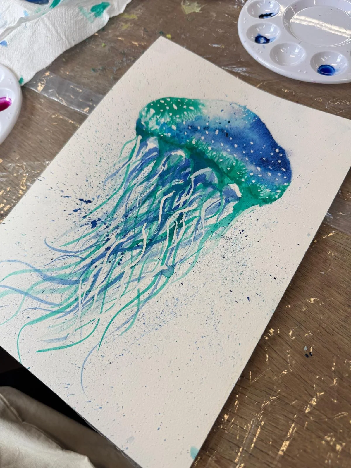 Watercolor painting of a jellyfish with blue, teal, and green hues, delicate tentacles, and white dots representing the bell's texture.