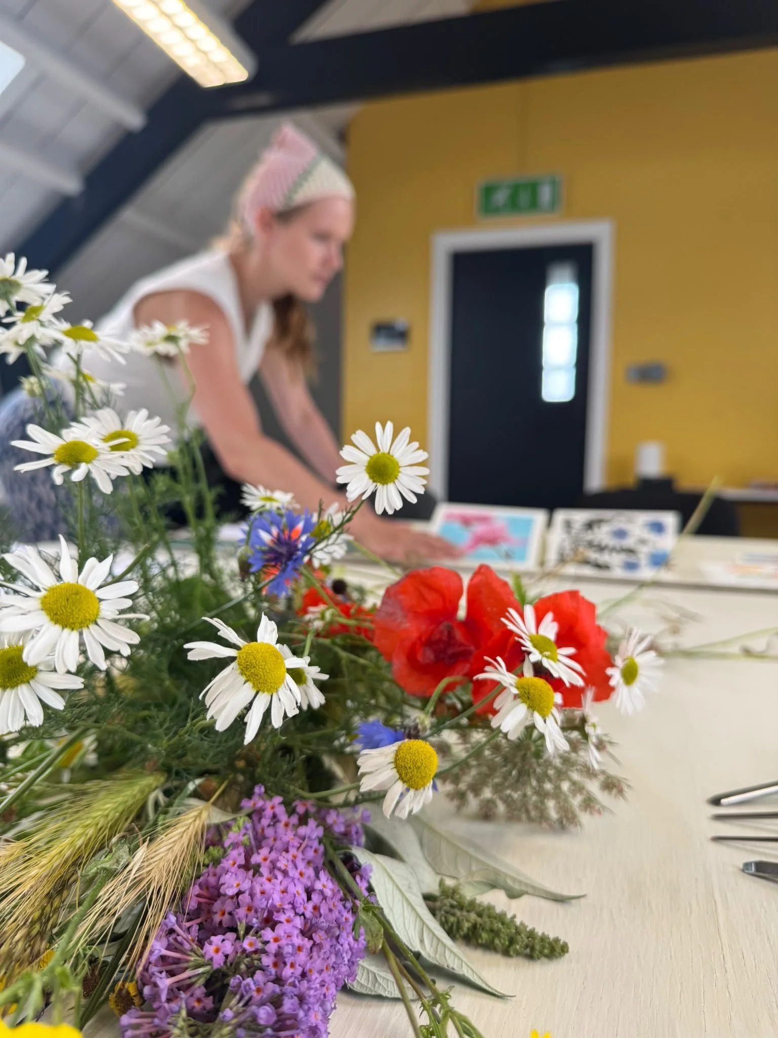Close-up of a colorful flower arrangement on a table with a blurred woman in the background, wearing a pink and white striped hat and working on something.