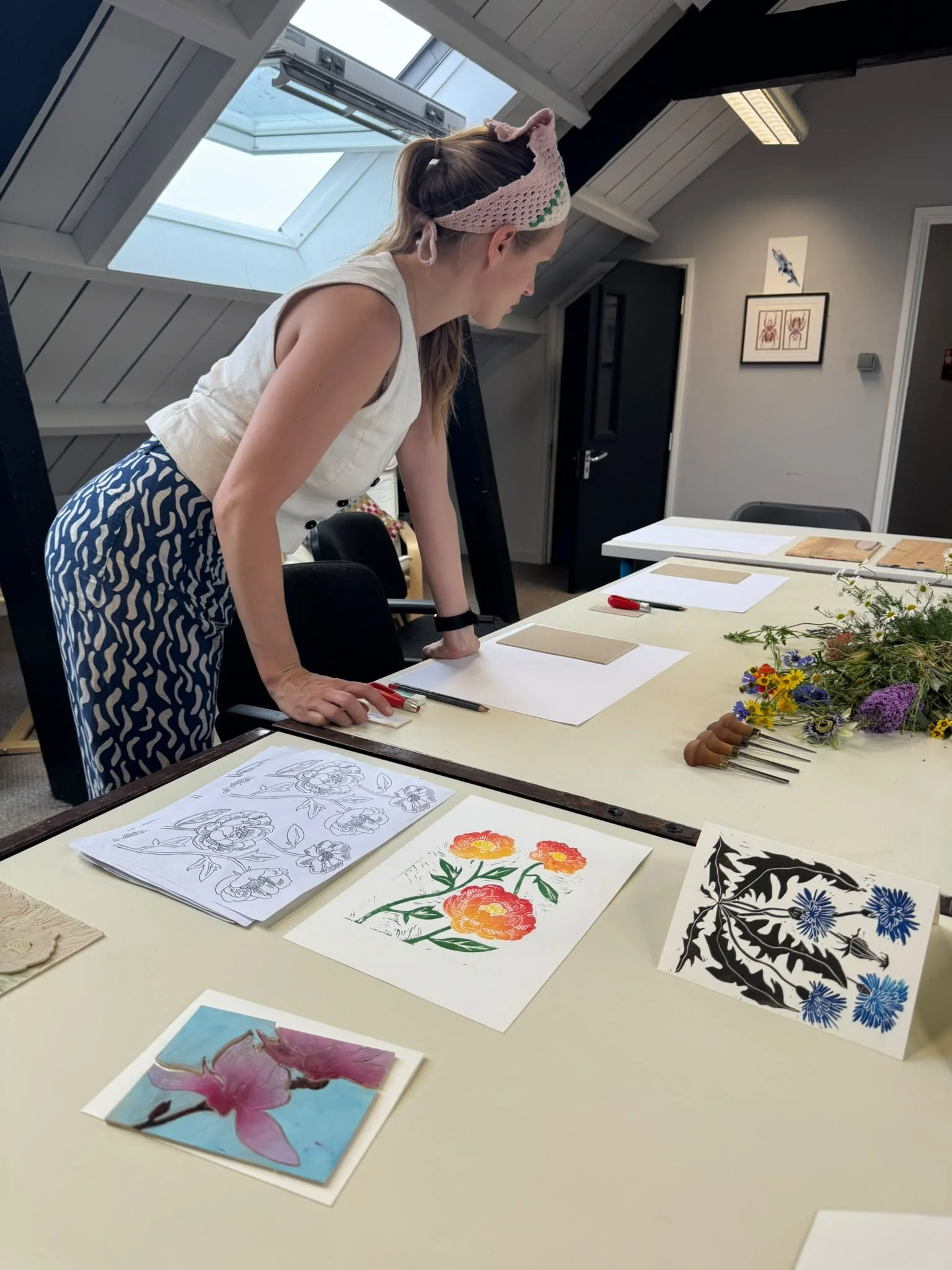 A woman in a sleeveless white top and patterned blue pants, wearing a knitted pink headband, stands in a room with sloped ceilings and skylights. She is leaning over a table with various floral artwork, sketches, and printing plates, while flowers an