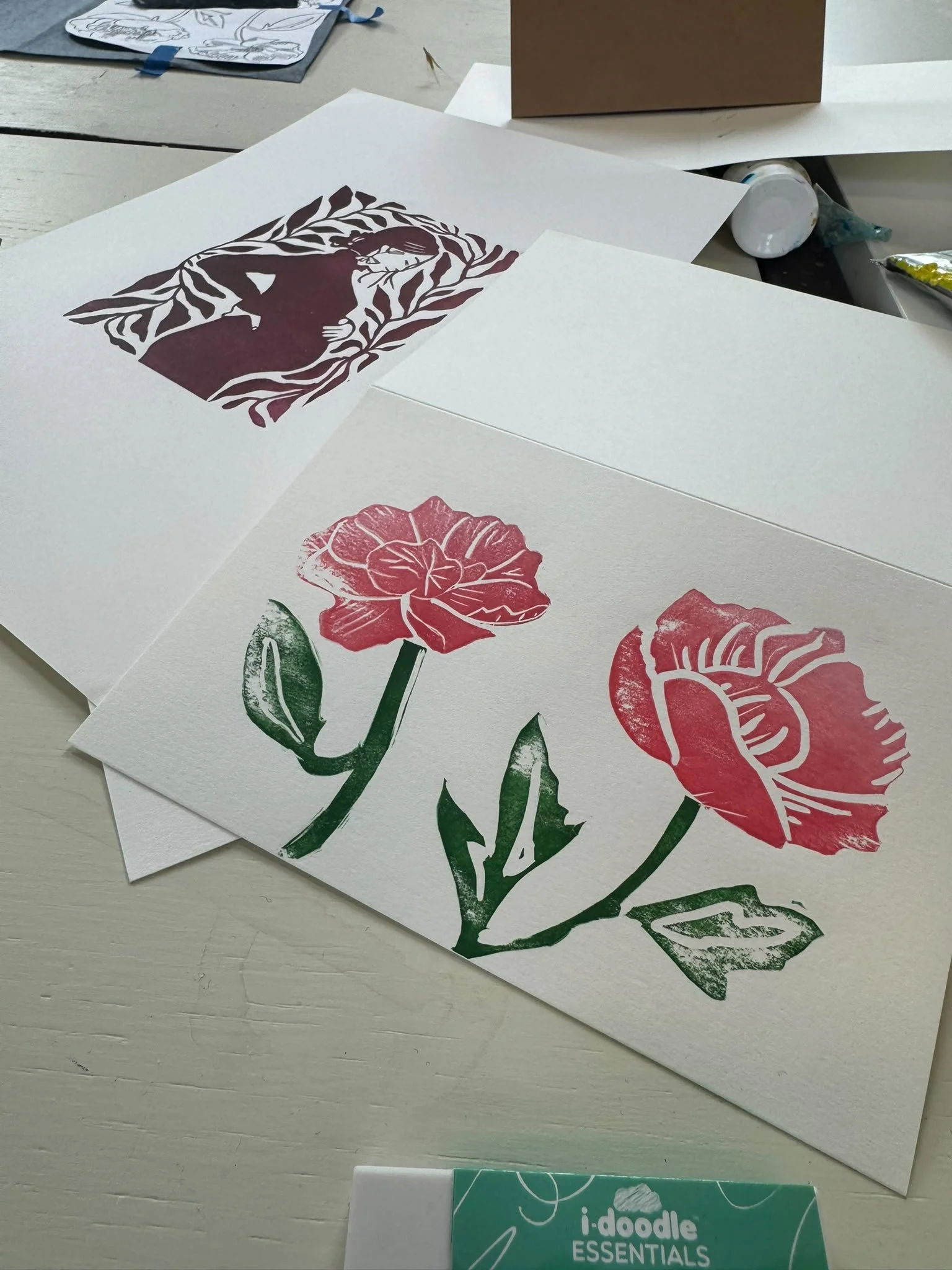 Paper prints of floral designs, including a purple flower with green leaves and a red flower with green leaves, on a light-colored table, with art supplies nearby.