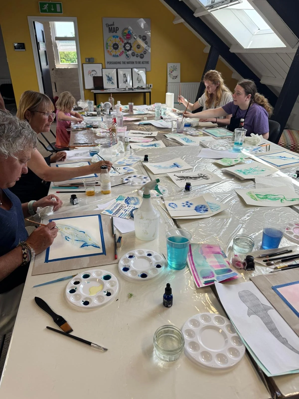People participating in a watercolor painting class in a room with a yellow wall and skylight windows. The table is covered with art supplies, paintings, and water glasses.