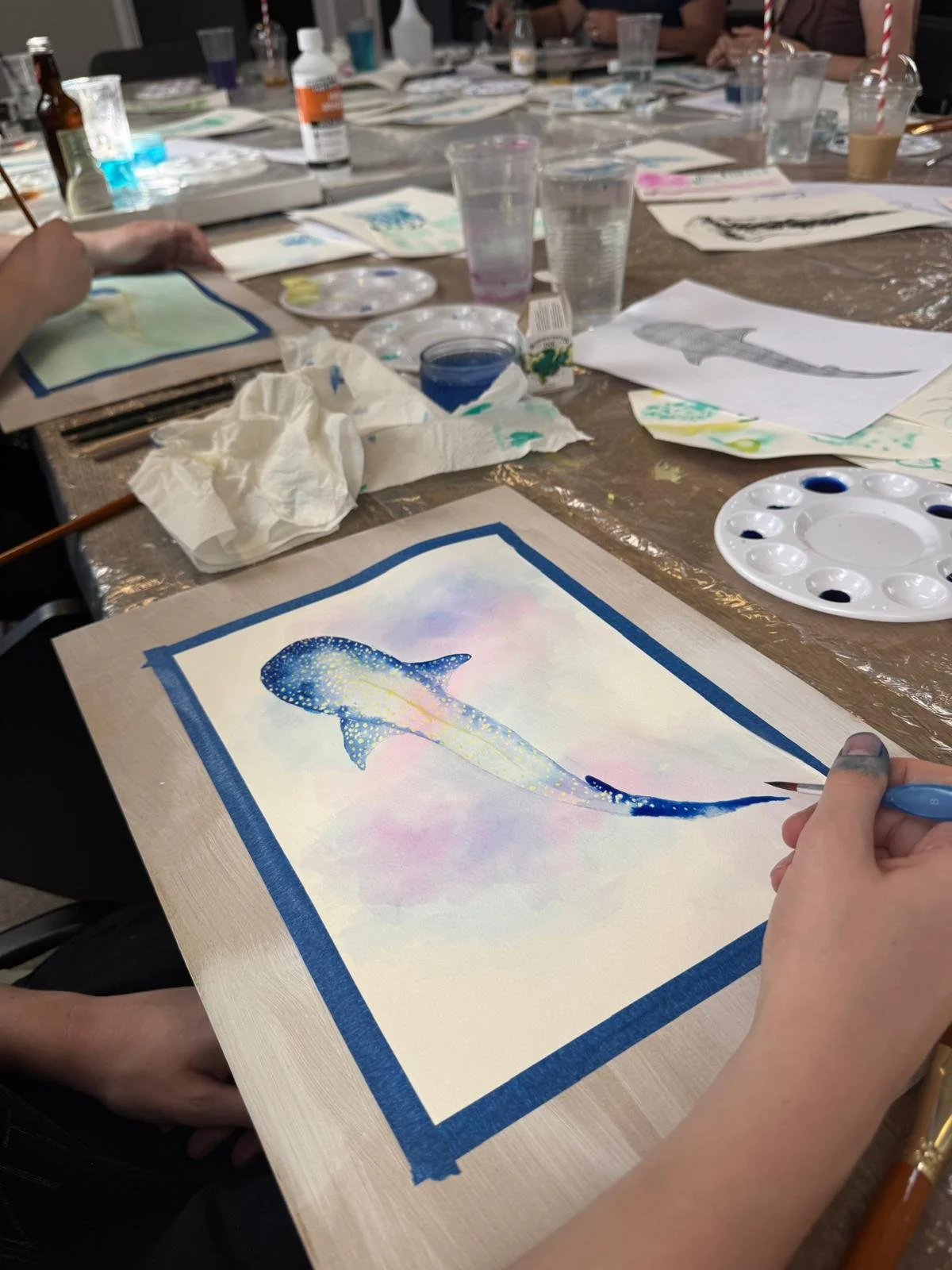 Person creating watercolor painting of a whale on paper at an art workshop, with various painting supplies and partially painted watercolors on the table.