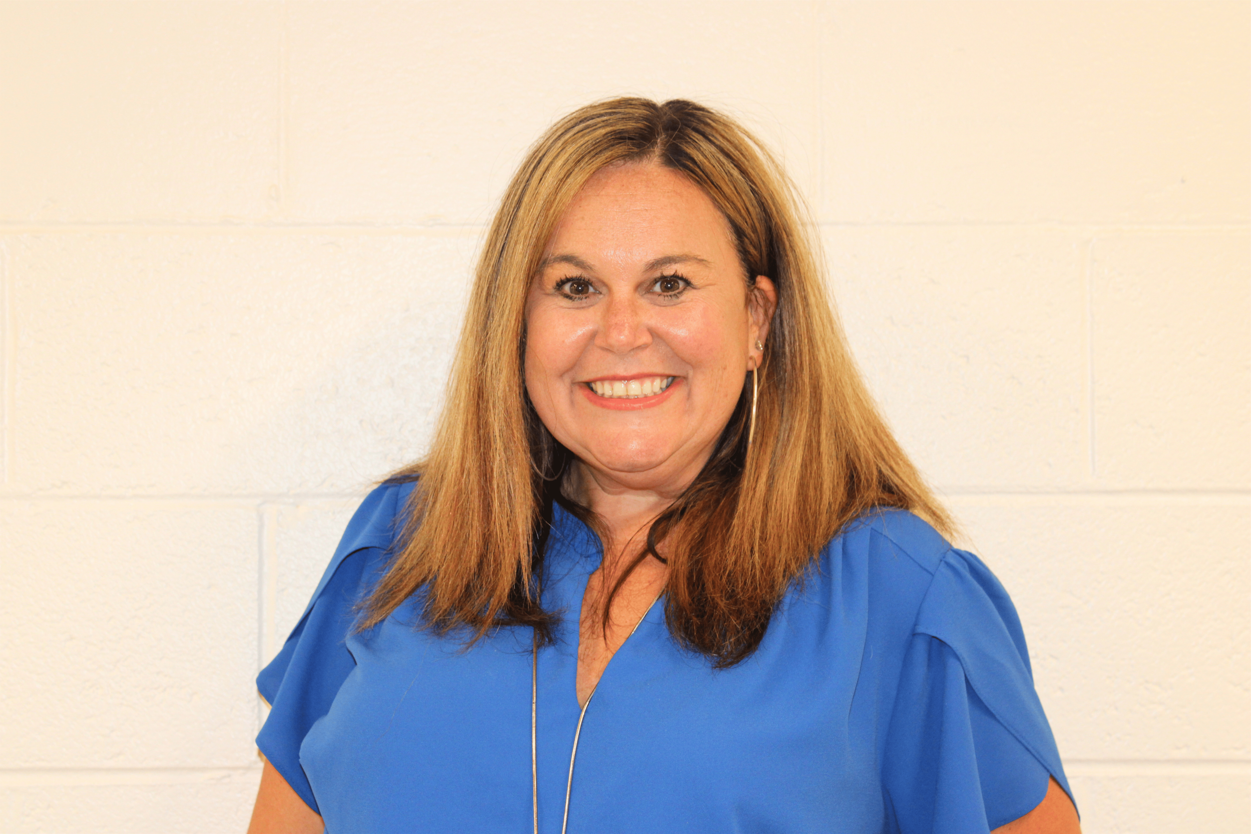 A woman with long, light brown hair and a big smile, wearing a blue top, standing against a white brick wall.