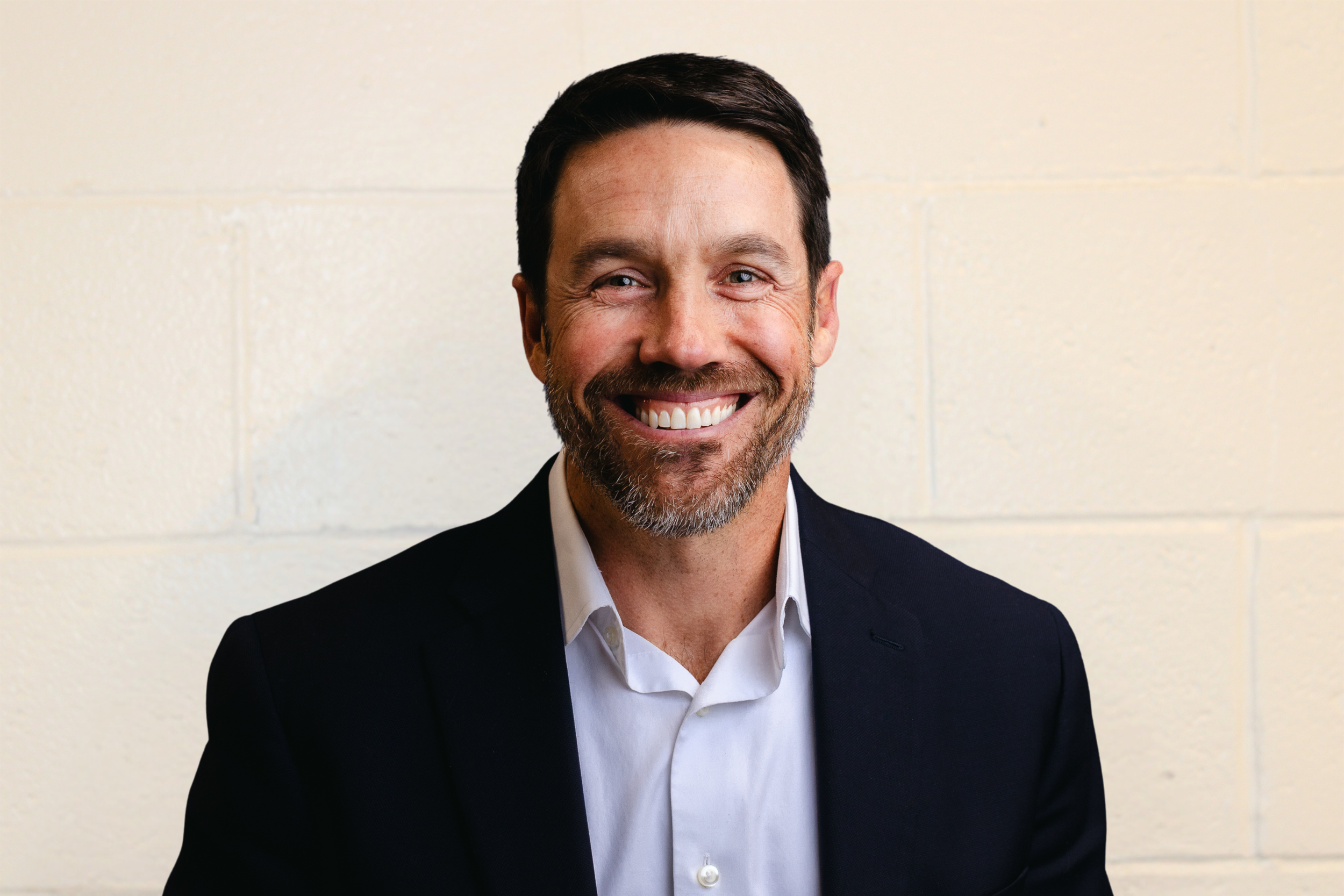 Portrait of a smiling man with dark hair and a beard, wearing a black blazer and white shirt, standing against a light-colored brick wall.