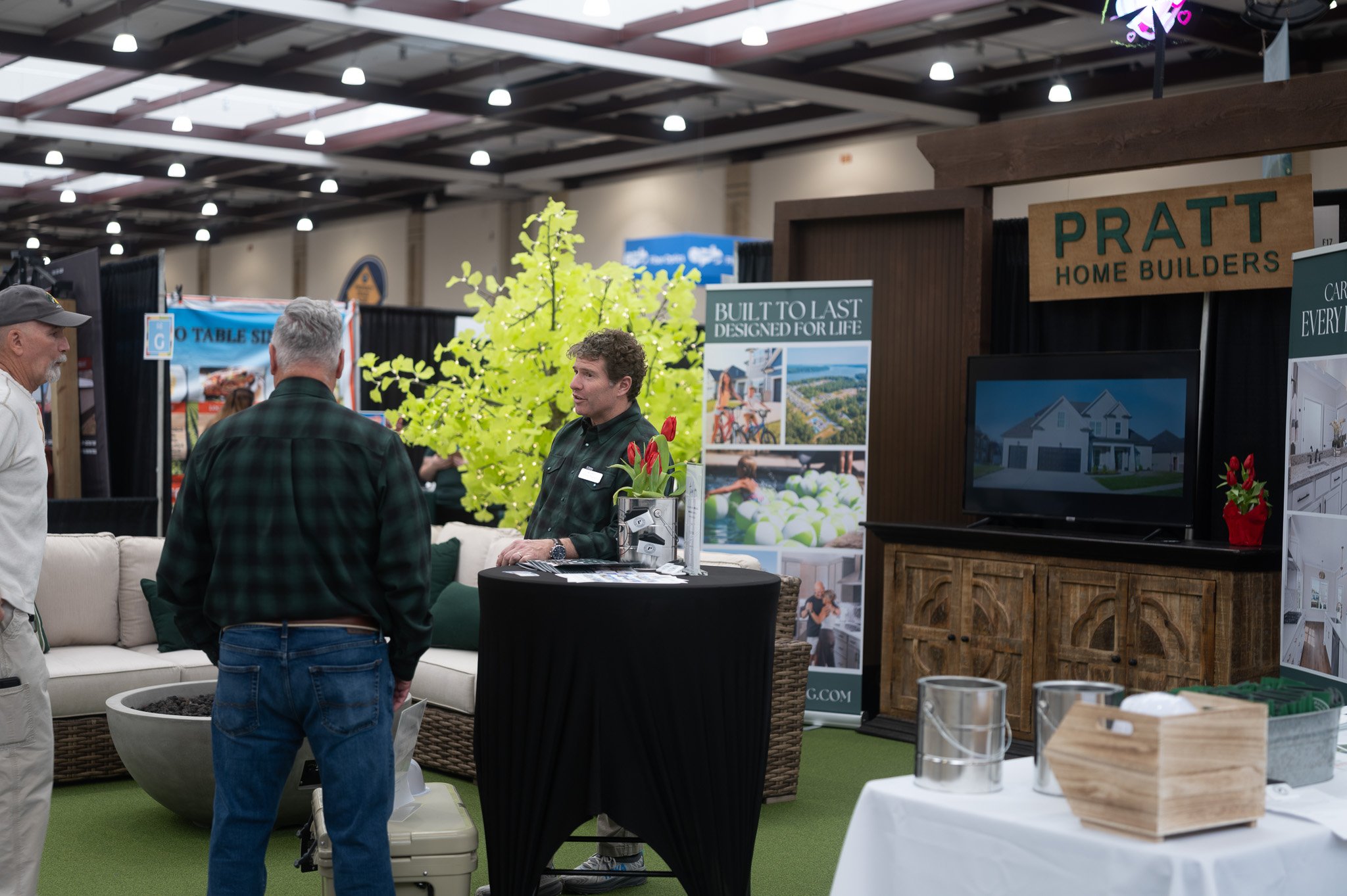 People attending a home building exhibition with booths for Pratt Home Builders; a display with a sign saying 'Built to Last Designed for Life' and pictures of houses, surrounded by plants and furniture.