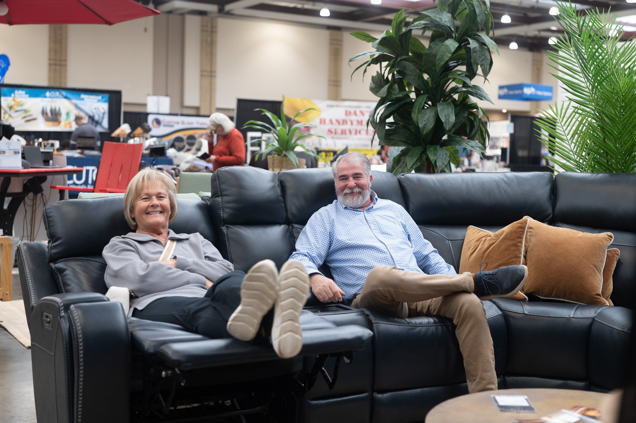 A smiling older woman and man sitting on a black leather couch in what appears to be a furniture store or indoor market, surrounded by large green plants.