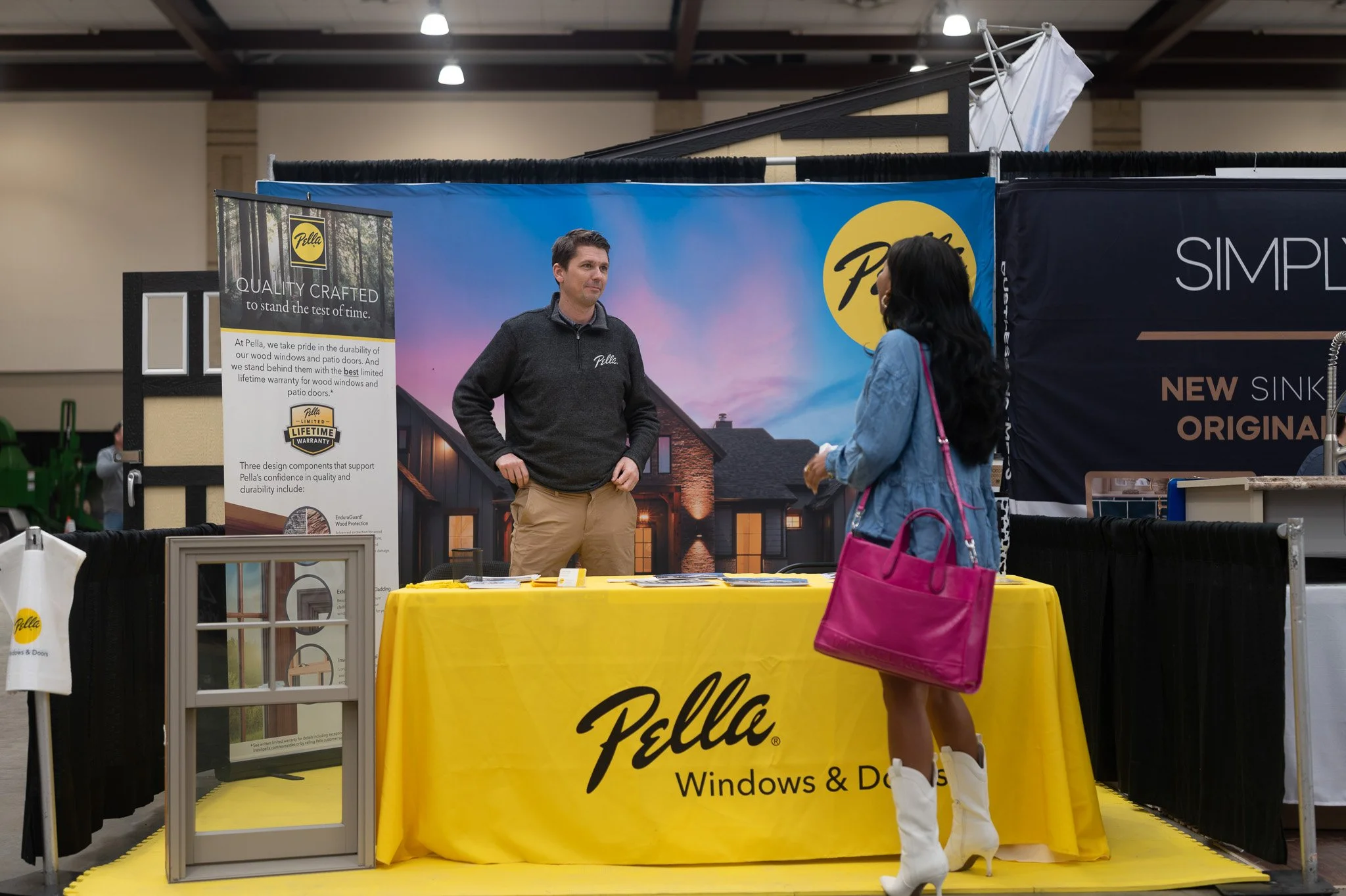 A man standing behind a booth with a yellow tablecloth displaying the Pella logo, speaking with a woman in a denim jacket carrying a pink purse, at an indoor exhibition or trade show. The booth features promotional materials for Pella Windows & Doors