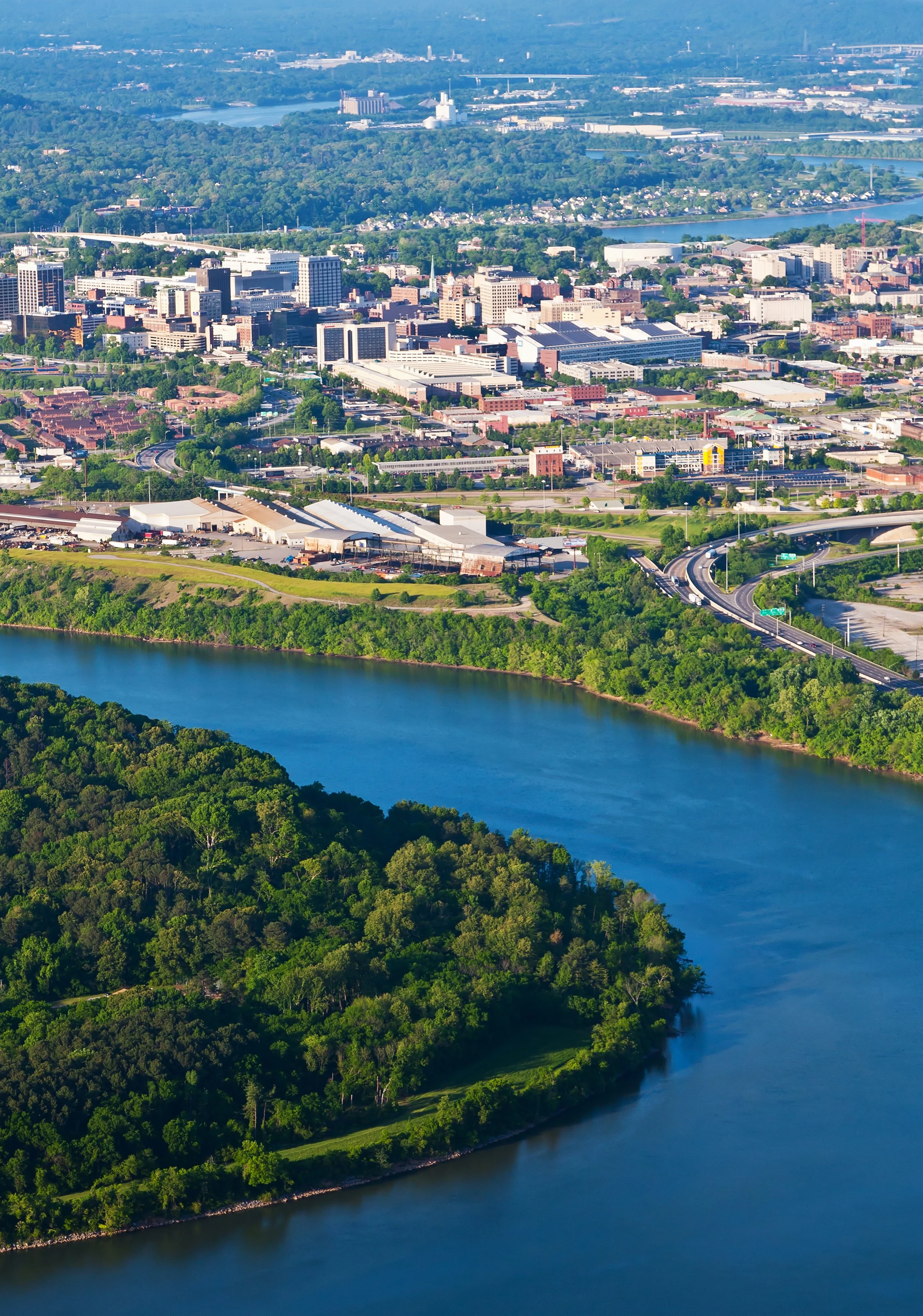 Aerial view of a city with tall buildings, highways, and surrounded by green forests and a river.