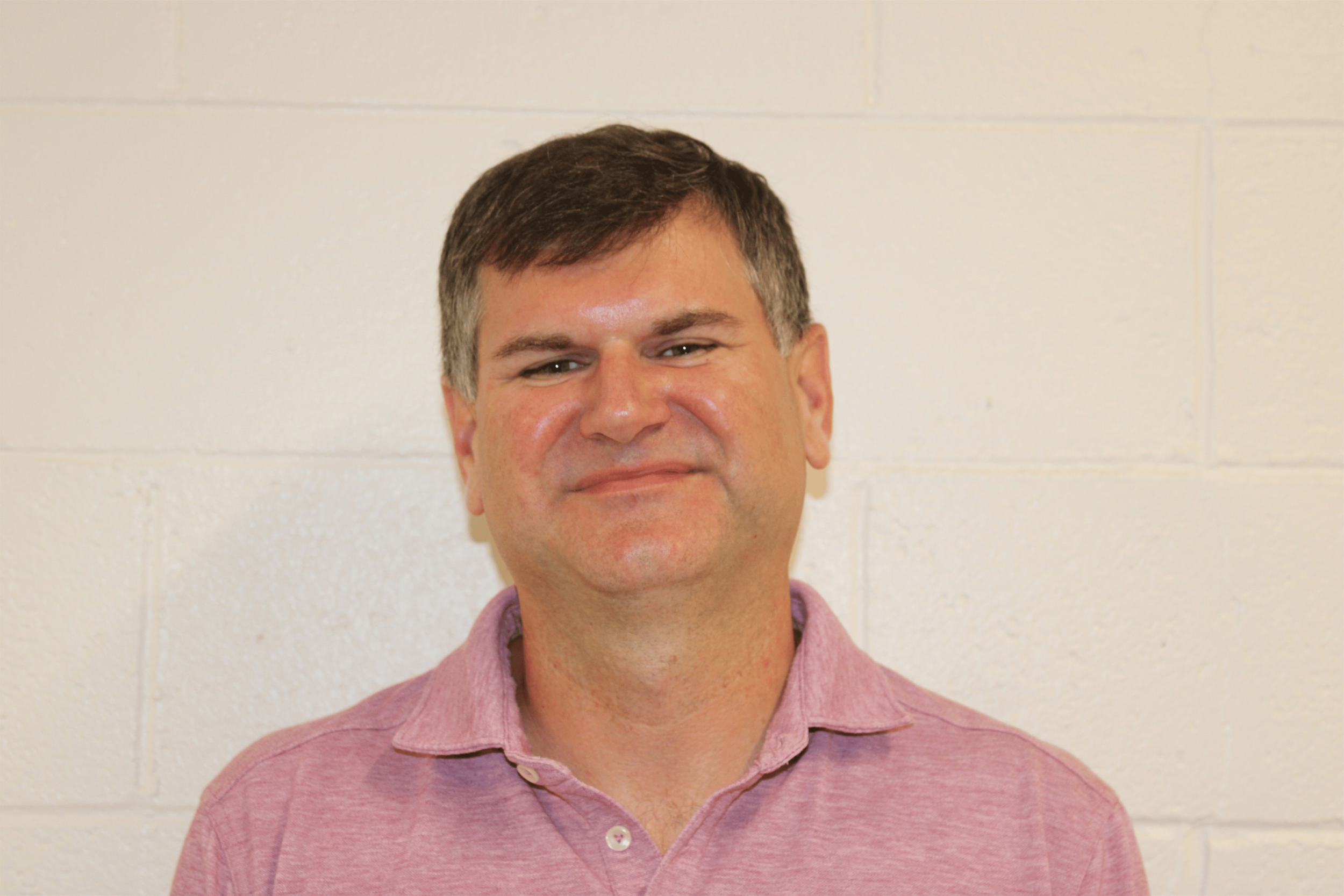 A man with short brown hair wearing a pink collared shirt stands in front of a white brick wall.