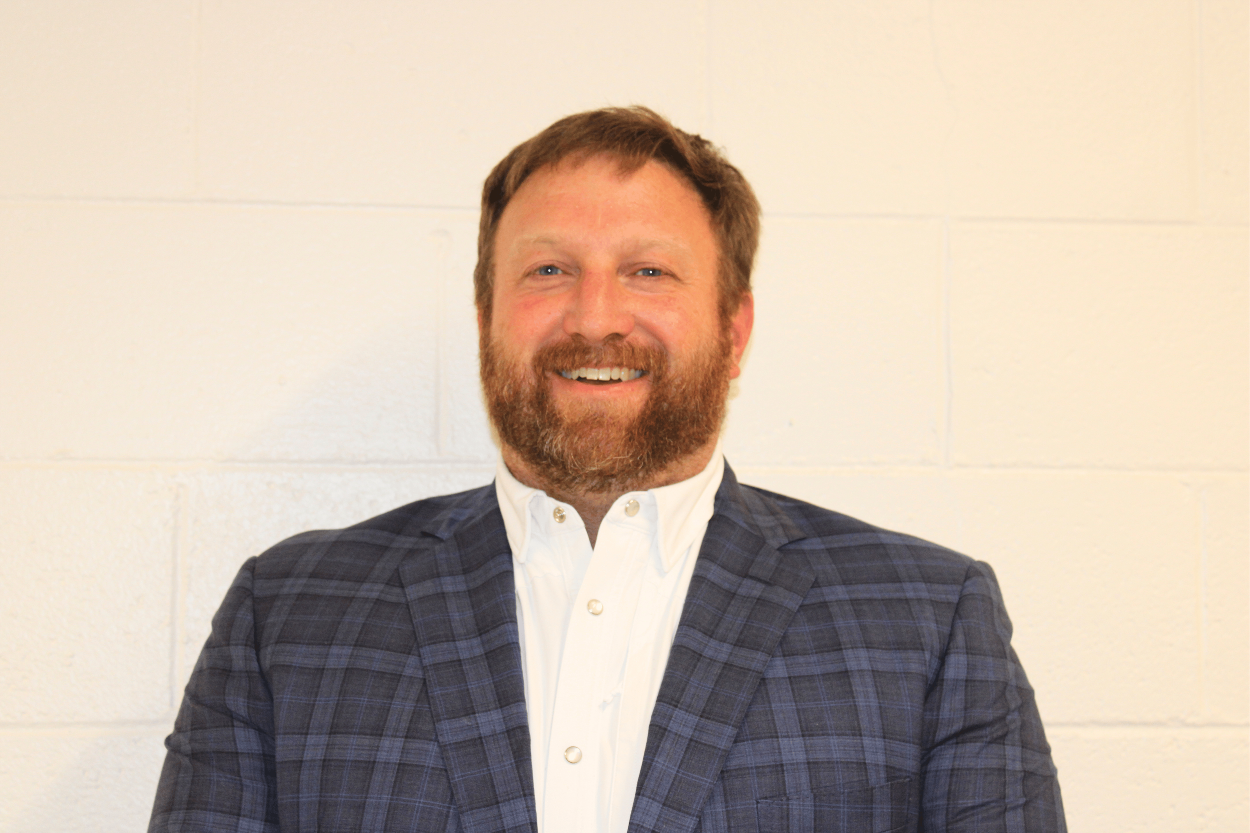 A smiling man with a beard and blue eyes, wearing a white shirt and a dark blue plaid blazer, standing against a beige wall.