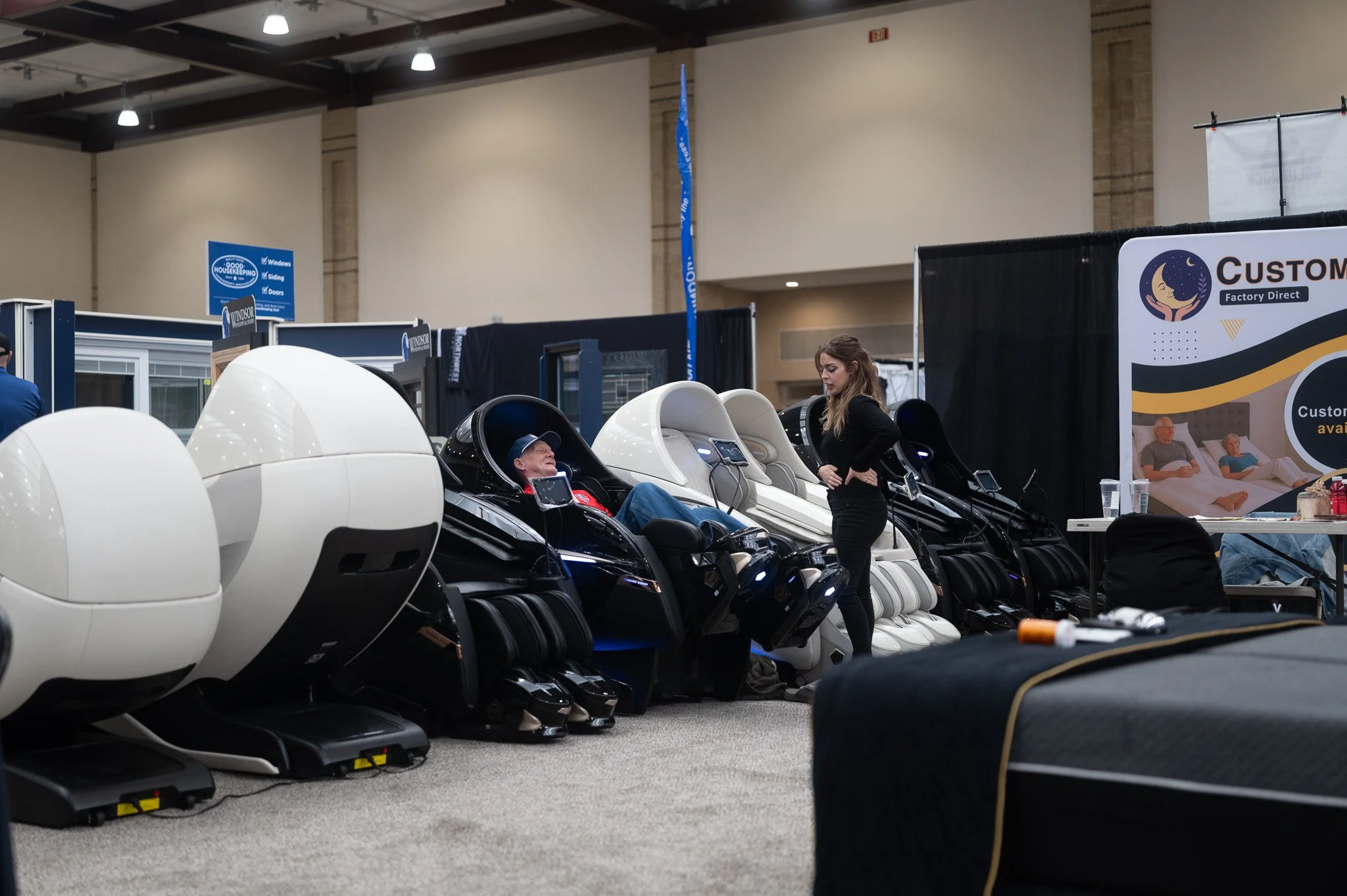 A woman demonstrating a massage chair to a man at an indoor trade show. Several massage chairs are displayed in a row, and a large sign with promotional information is visible in the background.