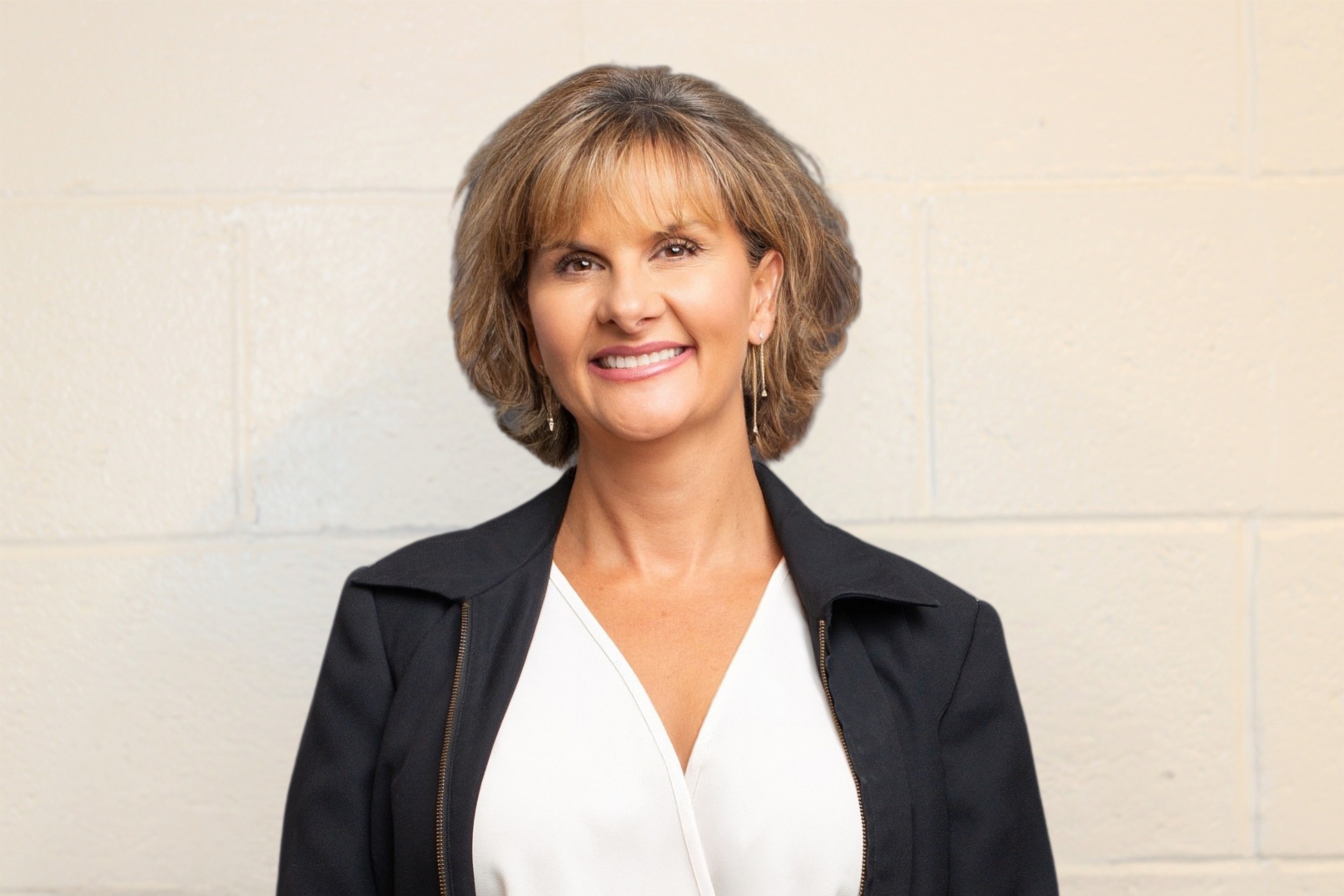 Professional woman with short, wavy blonde hair wearing a black jacket and white top, standing against a plain beige wall.