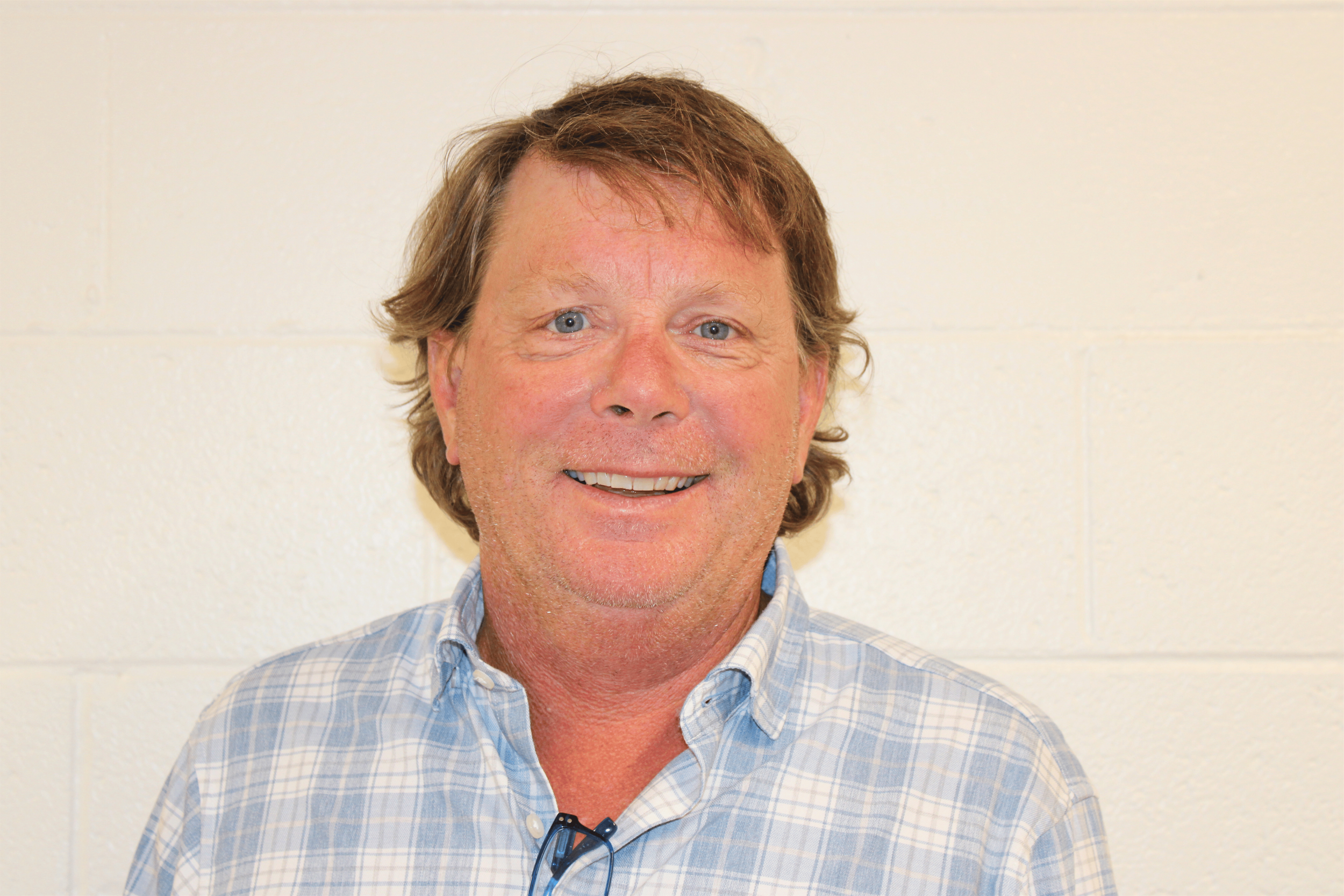 A middle-aged man with light brown, slightly wavy hair, blue eyes, and fair skin smiling at the camera. He is wearing a blue and white plaid shirt and has black glasses hanging from his shirt collar. The background is a plain, off-white wall.