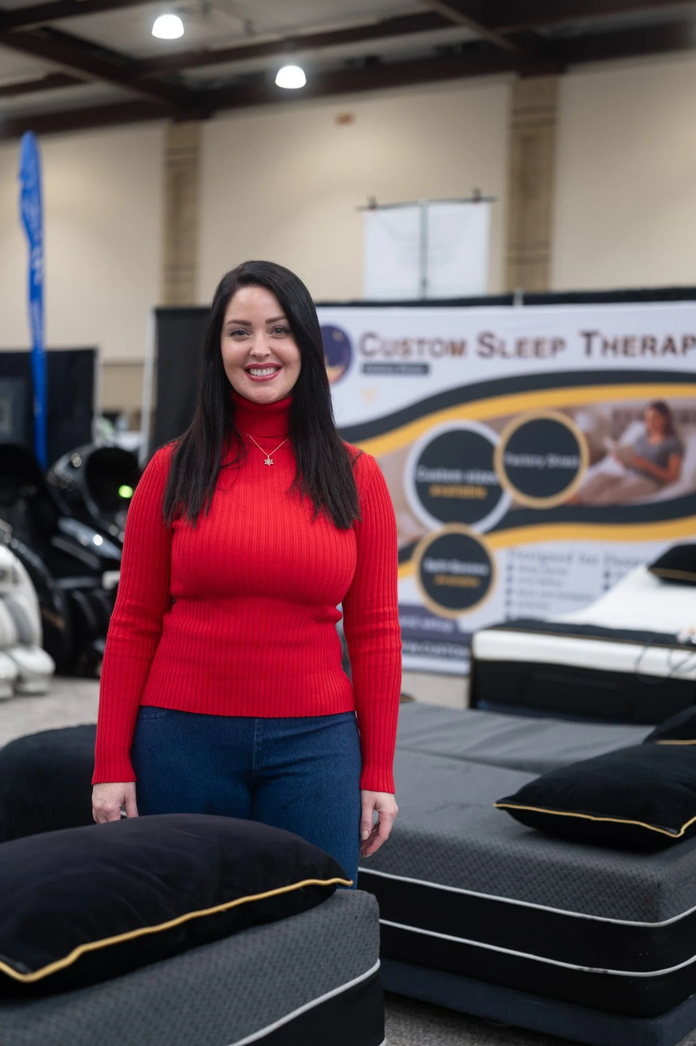A woman with long dark hair smiling, wearing a red turtleneck sweater and blue jeans, standing in an indoor showroom with beds and mattresses on display, and a banner in the background reading 'Custom Sleep Therapy'.